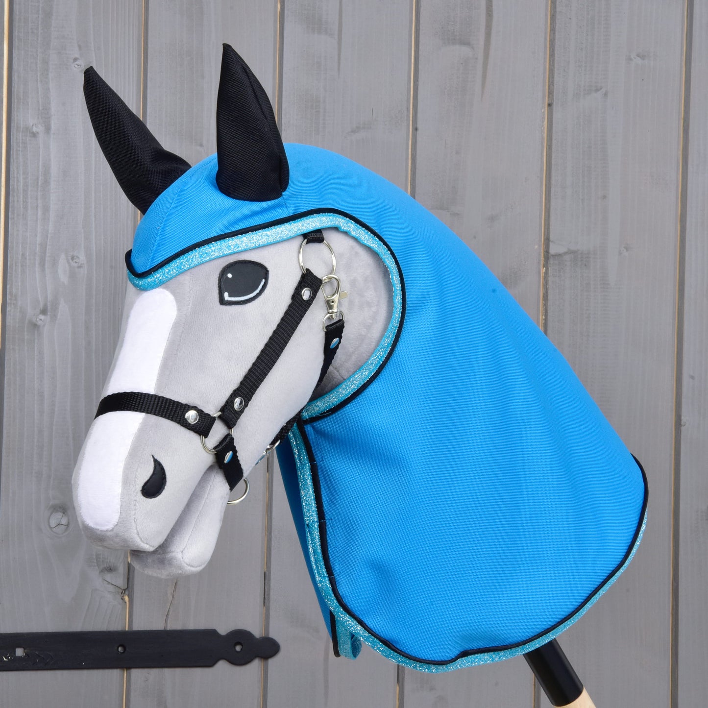 Imperméable Bleu Ciel de Hobby Horse LarDen, avec un Hobby Horse gris et blanc aux rênes noires et aux oreilles noires, portant une capuche brillante style imperméable bleu ciel, posé devant un fond en bois.