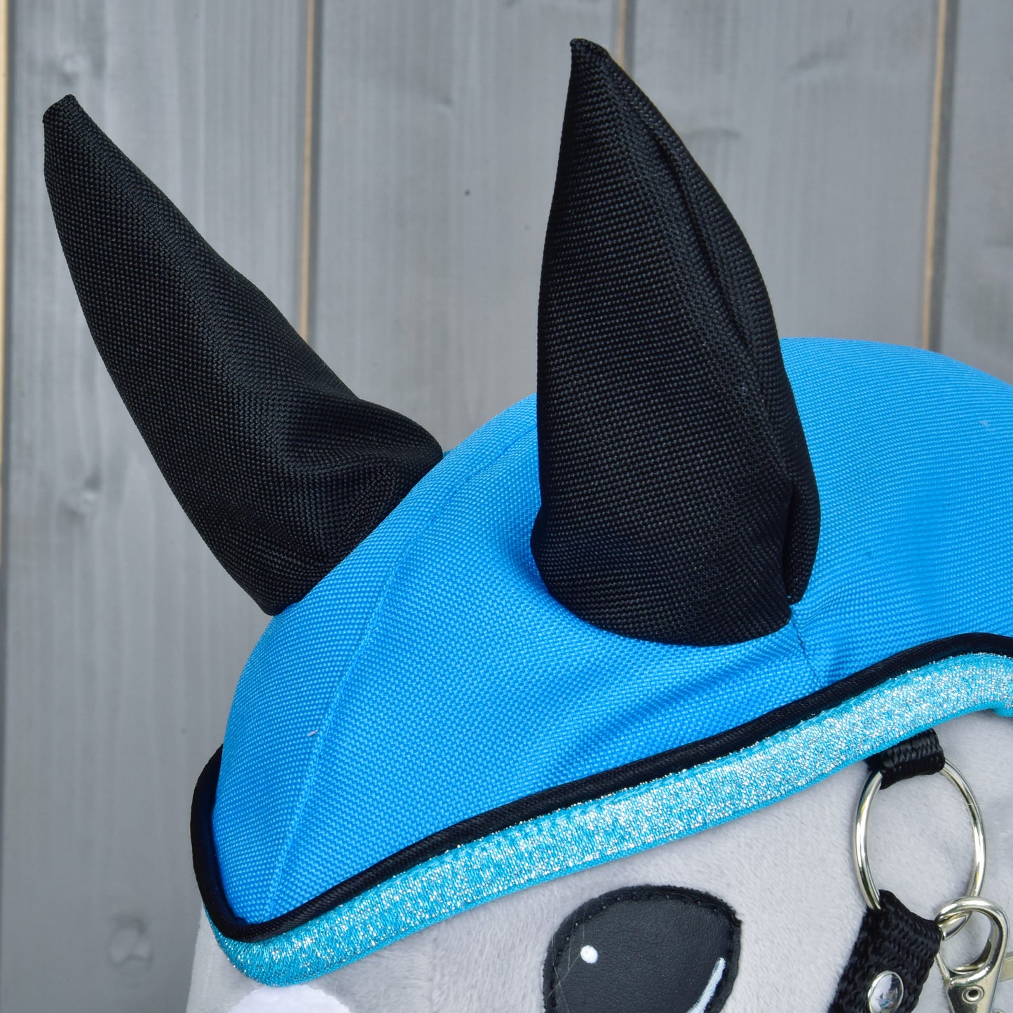 Gros plan sur une peluche Hobby Horse LarDen portant l'Imperméable Bleu Ciel, avec oreilles noires pointues et bordure bleue scintillante. L'imperméable par tous les temps ressort sur un fond en panneaux de bois gris clair.