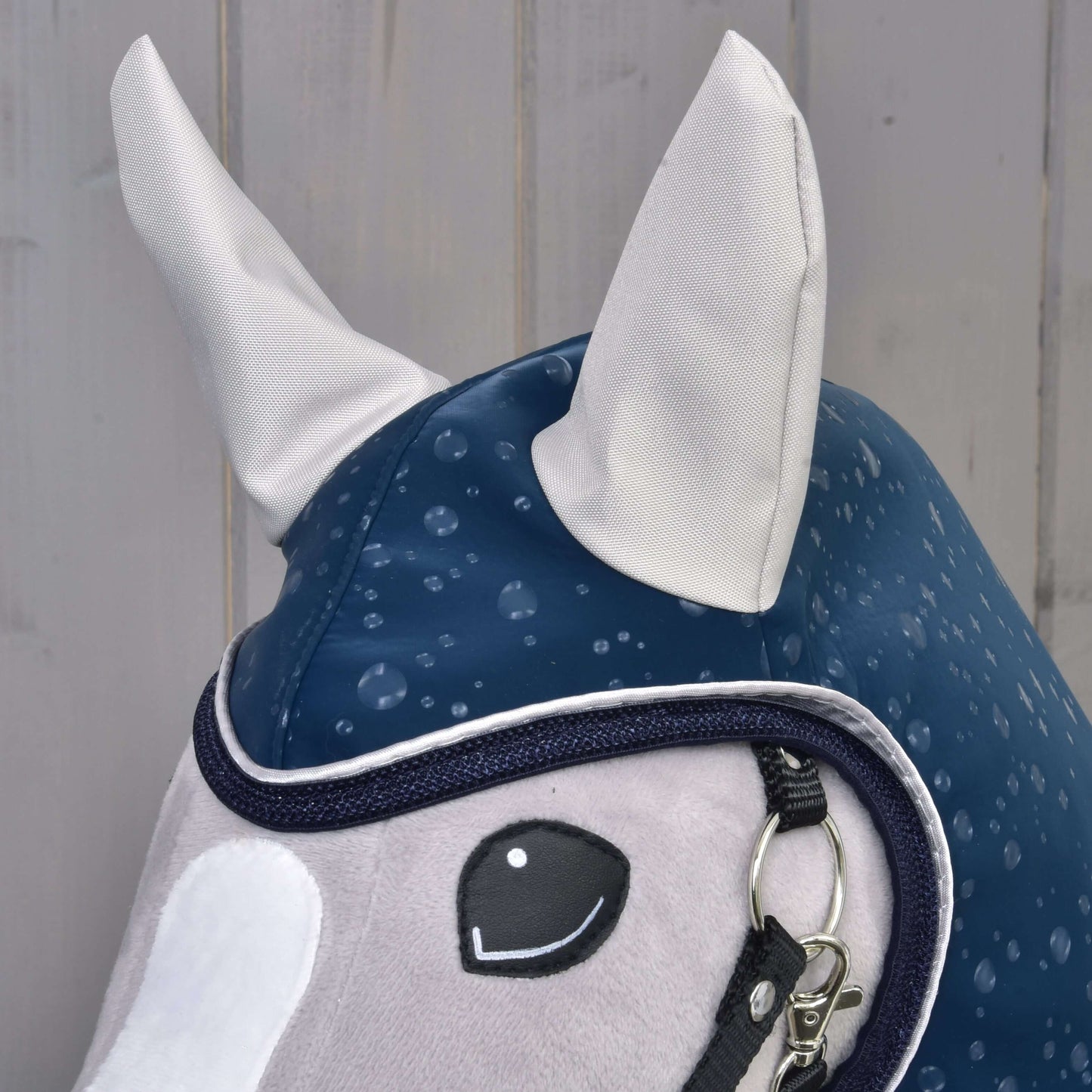 Gros plan sur le Hobby Horse LarDen "Raincoat Derby" en peluche, avec visage gris clair, oreilles blanches, rênes noires et capuche d’imperméable bleu marine pour tous les temps, sur fond en bois clair.