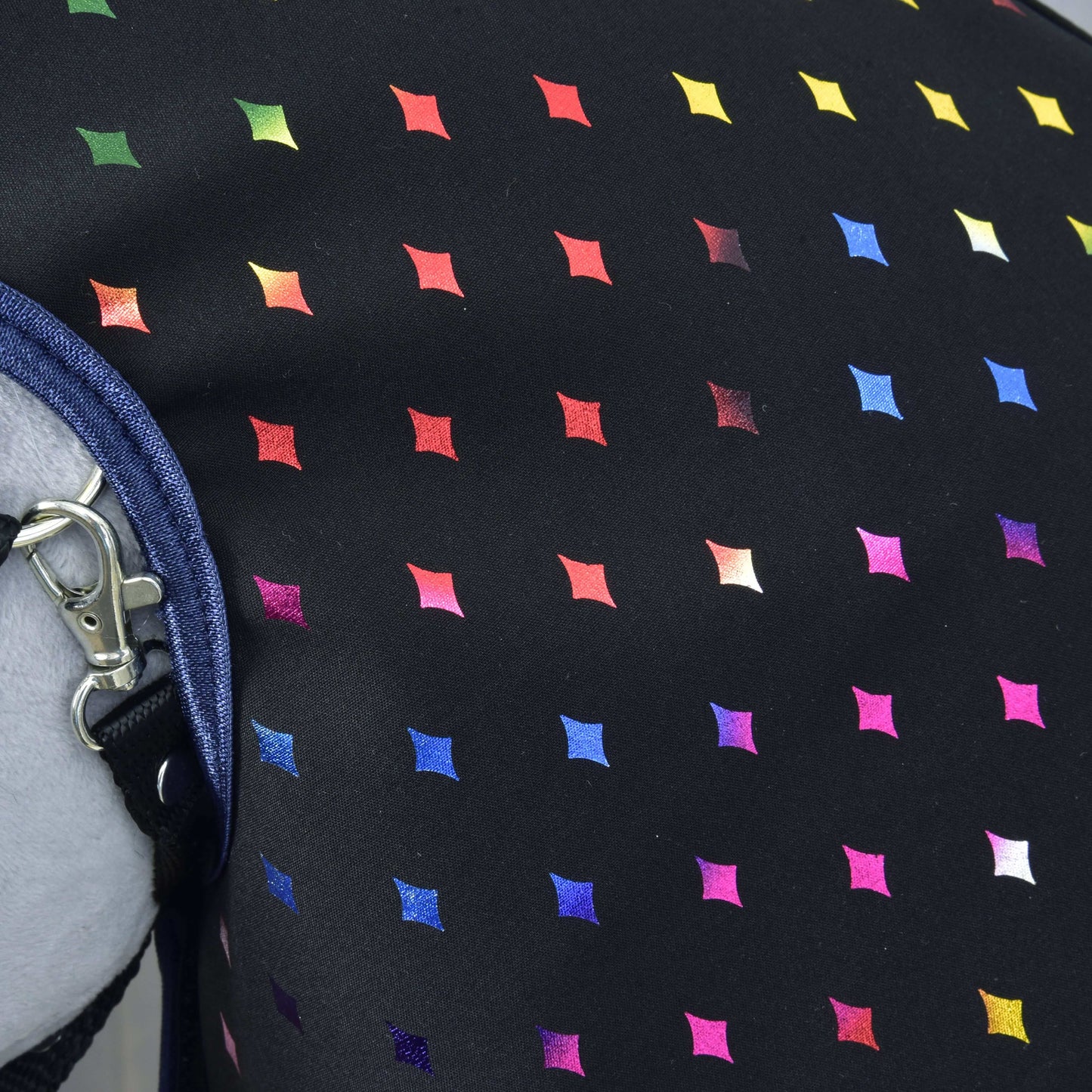 Gros plan sur l’Imperméable Reine Étincelante de Hobby Horse LarDen, avec tissu noir à grille de petits losanges colorés. Un fermoir métallique et une sangle en jean bleu à gauche soulignent sa polyvalence par tous les temps.