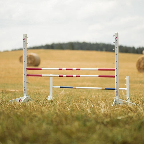 Weiß-rot gestreifte Sprungstangen auf einer grasbewachsenen Fläche, ideal als Hindernis für Hobby Horsing, mit Heuballen und Bäumen im Hintergrund unter bewölktem Himmel.
