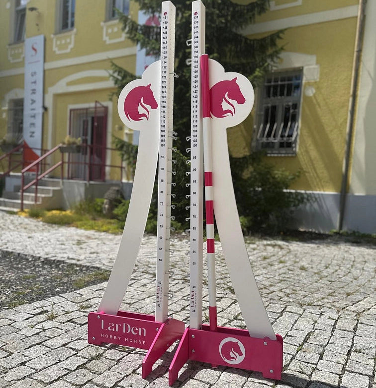 Zwei weiße und pinke verstellbare Sprungständer für Hobby Horsing stehen draußen auf einem Kopfsteinpflasterweg, vor einem gelben Gebäude mit Treppe und einem Schild im Hintergrund.