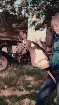 Lifestyle video: a young rider holds the Hobby Horse Monte by Hobby Horse LarDen in a meadow in front of an old wooden wagon - brown and white coat with white patches, long dark yarn mane, wooden stick, shown here with the fur-lined Espresso halter.