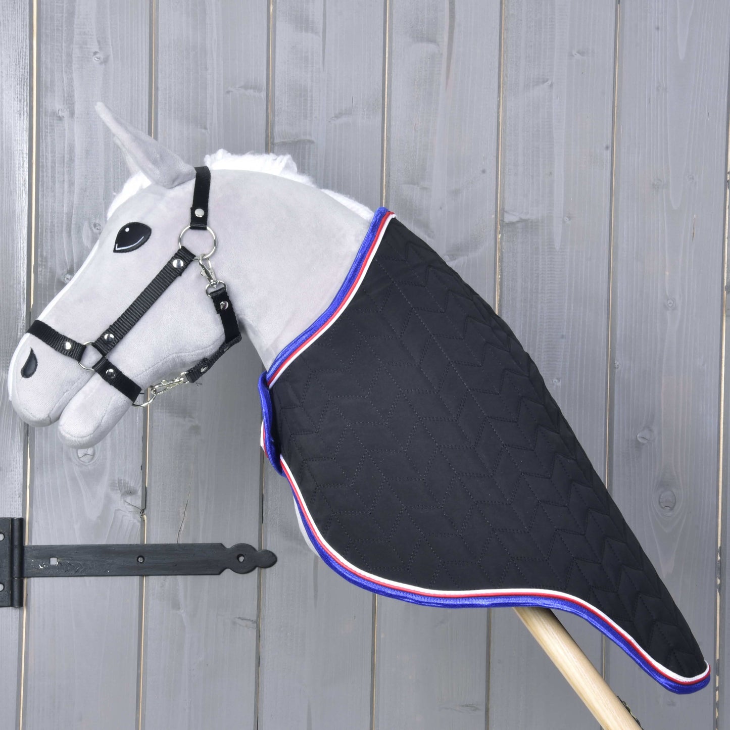 Couverture de voyage de luxe de Hobby Horse LarDen sur un jouet Hobby Horse gris monté sur un bâton en bois, couverture noire matelassée avec bordure rouge, blanche et bleue, sur fond rustique en bois.