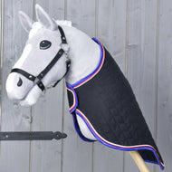 Ein graues Hobby Horse mit schwarzem Zaumzeug trägt die Hobby Horse LarDen Transportdecke Luxury – schwarz gesteppt und mit rot-weiß-blauer Borte – vor einer Holzwand.