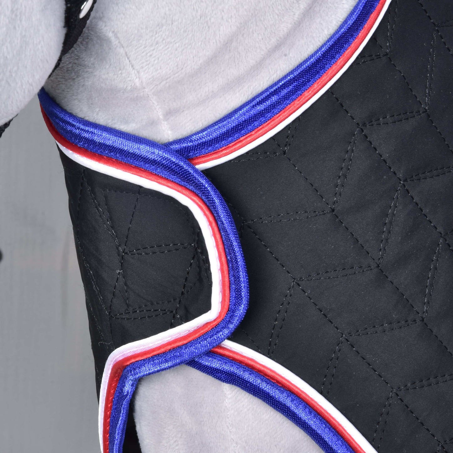 Gros plan sur un tissu noir matelassé avec bordure rouge, blanche et bleue, enveloppant une surface grise, probablement la Couverture de voyage de luxe de Hobby Horse LarDen, conçue pour un confort et une protection supérieurs pendant le transport.