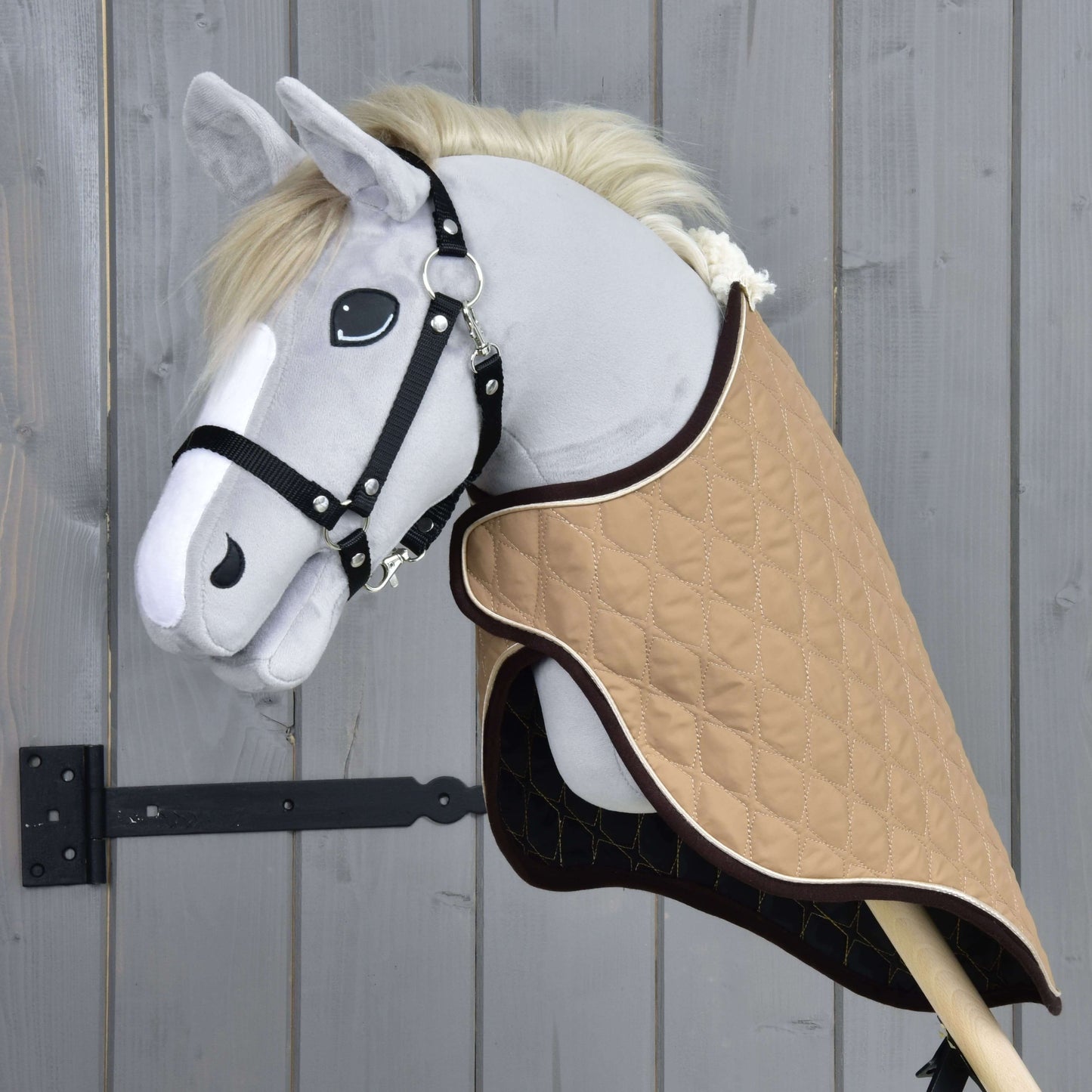 Ein graues Stoff-Hobby Horse von Hobby Horse LarDen mit schwarzem Zaumzeug und der Transportdecke Paris ist auf einem Holzstab montiert und vor einem Holzzaun platziert.