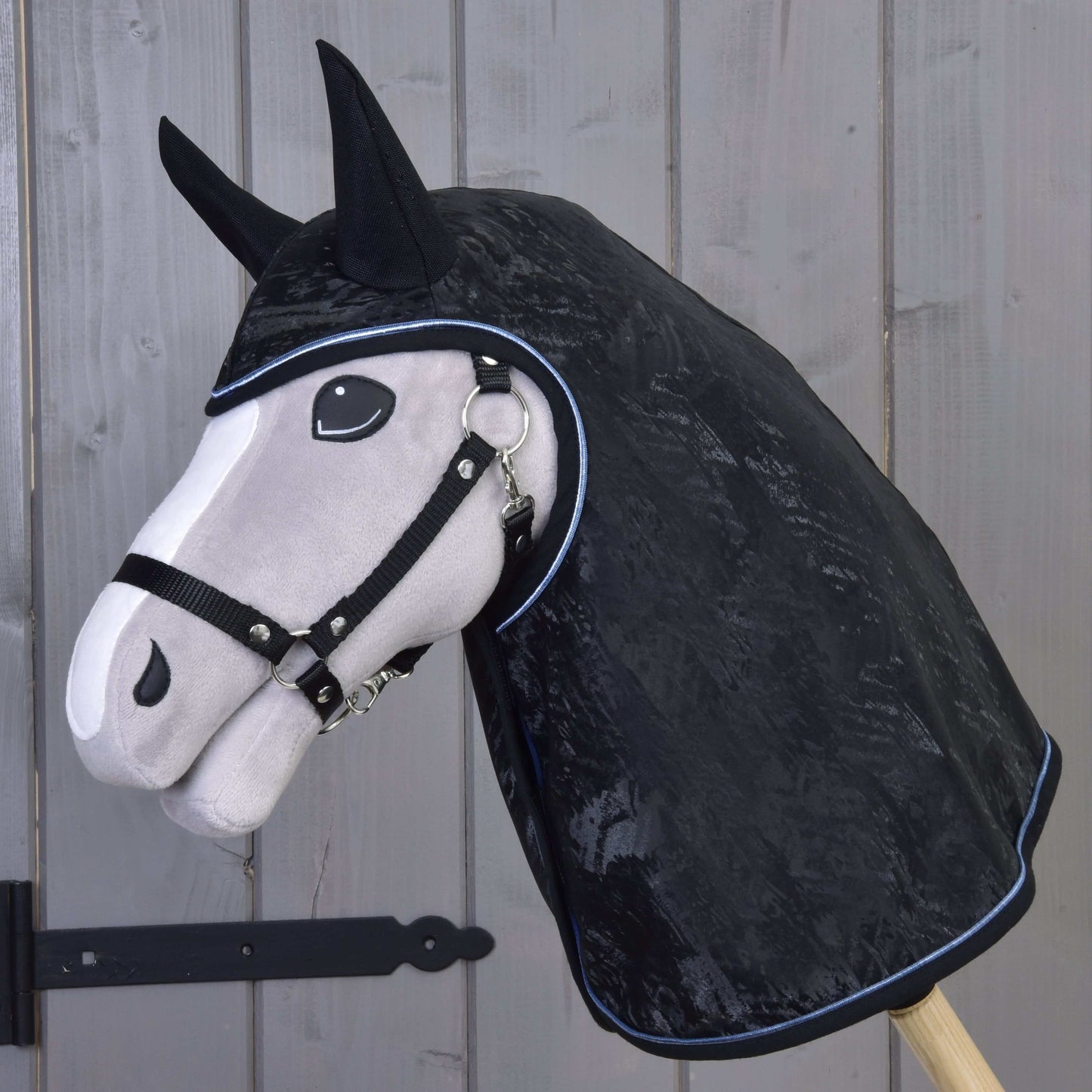Ein Stoffpferdekopf auf einem Holzstock trägt die Hobby Horse LarDen Regendecke Silver Stone mit Ohrenschutz und einem Halfter für Hobby Horse und steht vor einer holzvertäfelten Tür.
