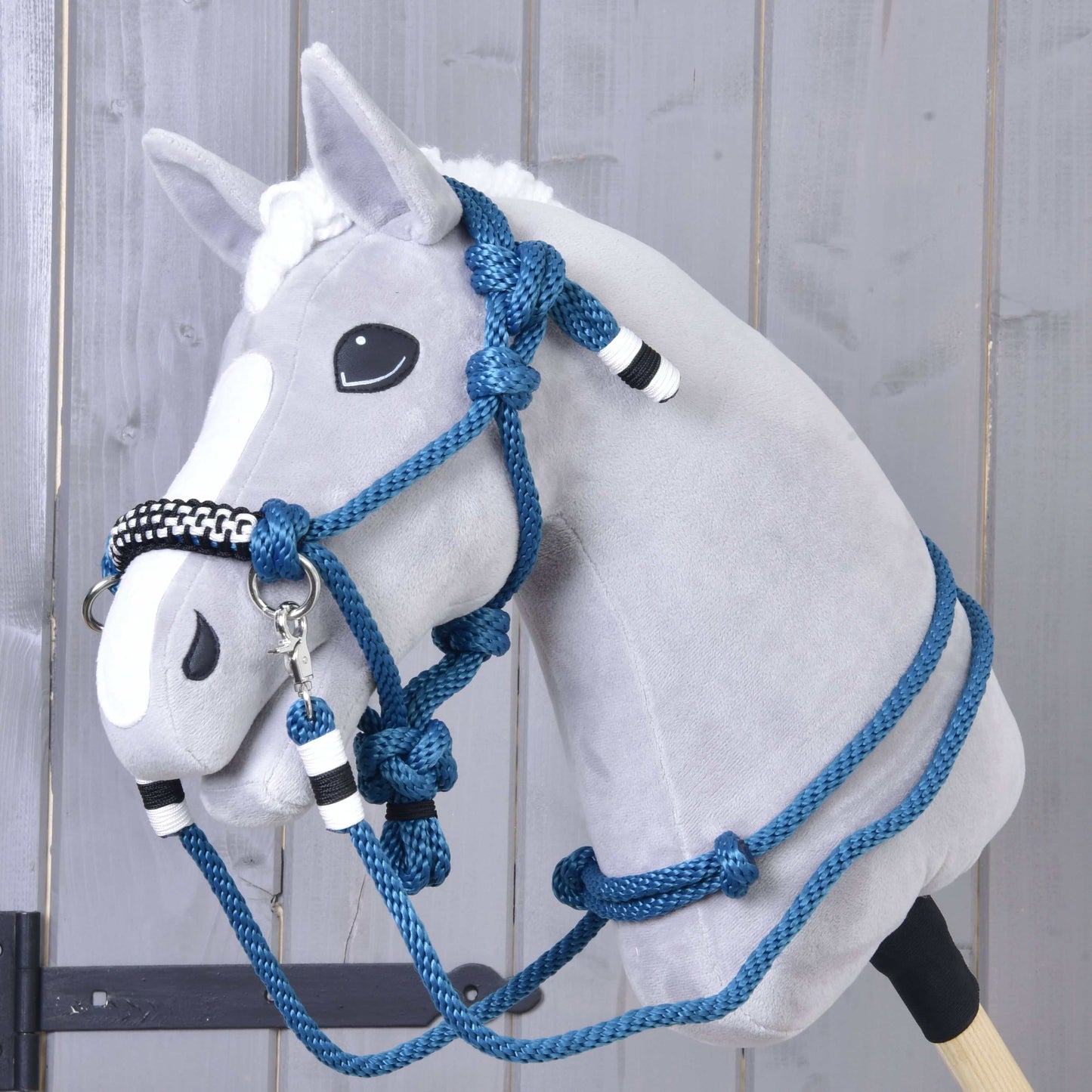 Seilset Chess von Hobby Horse LarDen an einem plüschigen Hobby Horse mit grauem Gesicht, weißer Blesse und blauen Halfter-Zügeln als Cordeo mit schwarzen und weißen Akzenten, vor einer Holz wand im Hintergrund.