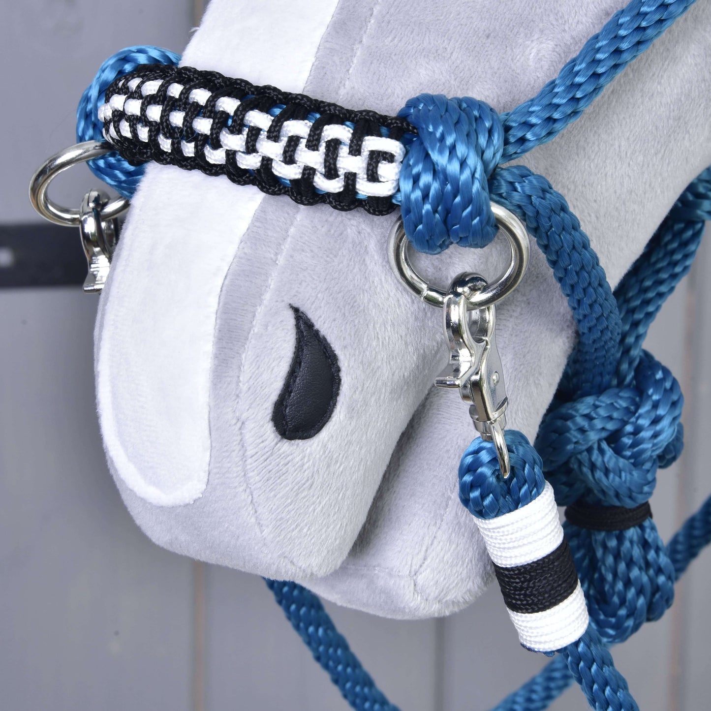 Nahaufnahme eines weichen grauen Pferdekopfs mit aufgesticktem schwarzem Auge, der das Hobby Horse LarDen Seilset Chess trägt – ein handgefertigtes blaues, schwarzes und weißes Halfter mit Metallringen und Clips.