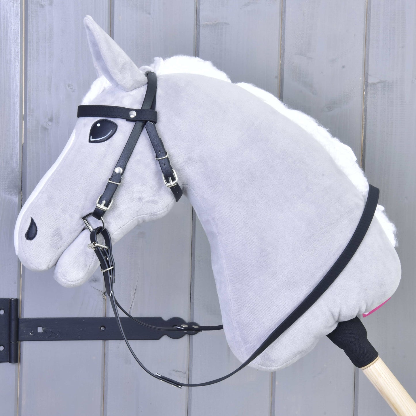 Nahaufnahme eines grauen Hobby Horse Spielzeugs von Hobby Horse LarDen mit Komplettes Zaumzeug ohne Nasenriemen Pure Black und abnehmbaren Zügeln, auf einem Holzstab montiert vor grauem Holzhintergrund.