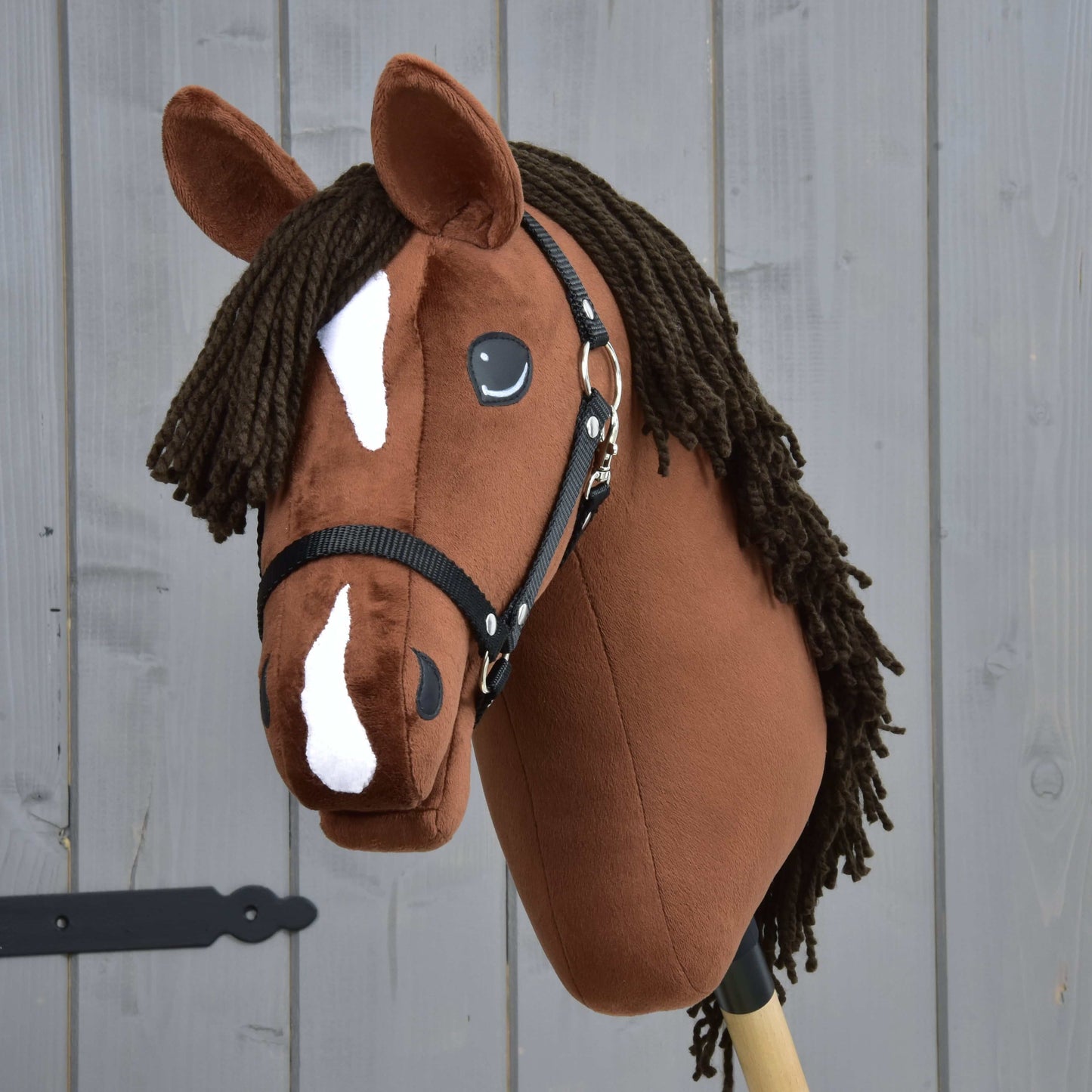 Hobby Horse Rocky avec licol noir de Hobby Horse LarDen avec tête marron de style pur-sang, marques blanches sur le visage et crinière foncée en fil, présenté sur fond de bois gris.