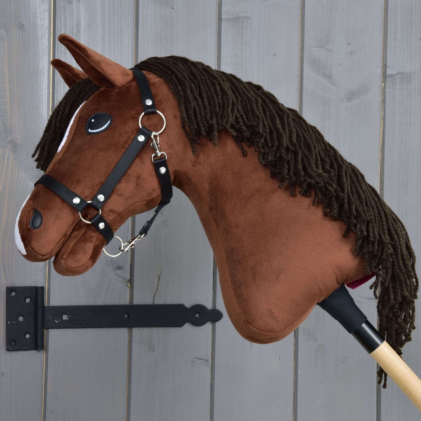 Hobby Horse Rocky avec licol noir de Hobby Horse LarDen, cheval-bâton marron en peluche, présenté à côté d’un mur en bois gris.