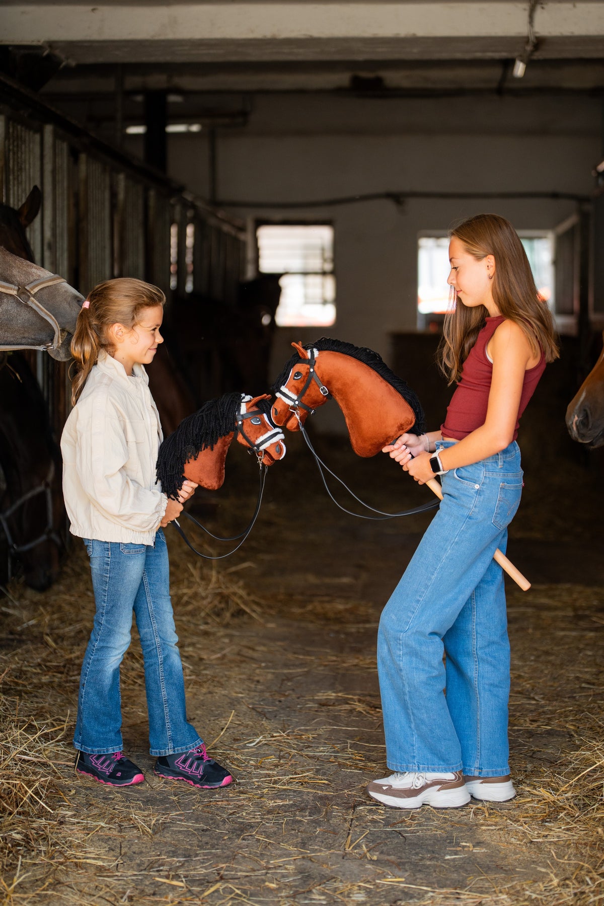 Zwei Mädchen stehen sich in einer Stallgasse gegenüber und halten jeweils ein Hobby Horse Rocky von Hobby Horse LarDen – fuchsbrauner Plüschkopf mit weißer Blesse und schwarzer Garnmähne – mit komplettem schwarzem Zaumzeug mit Fell und Zügeln