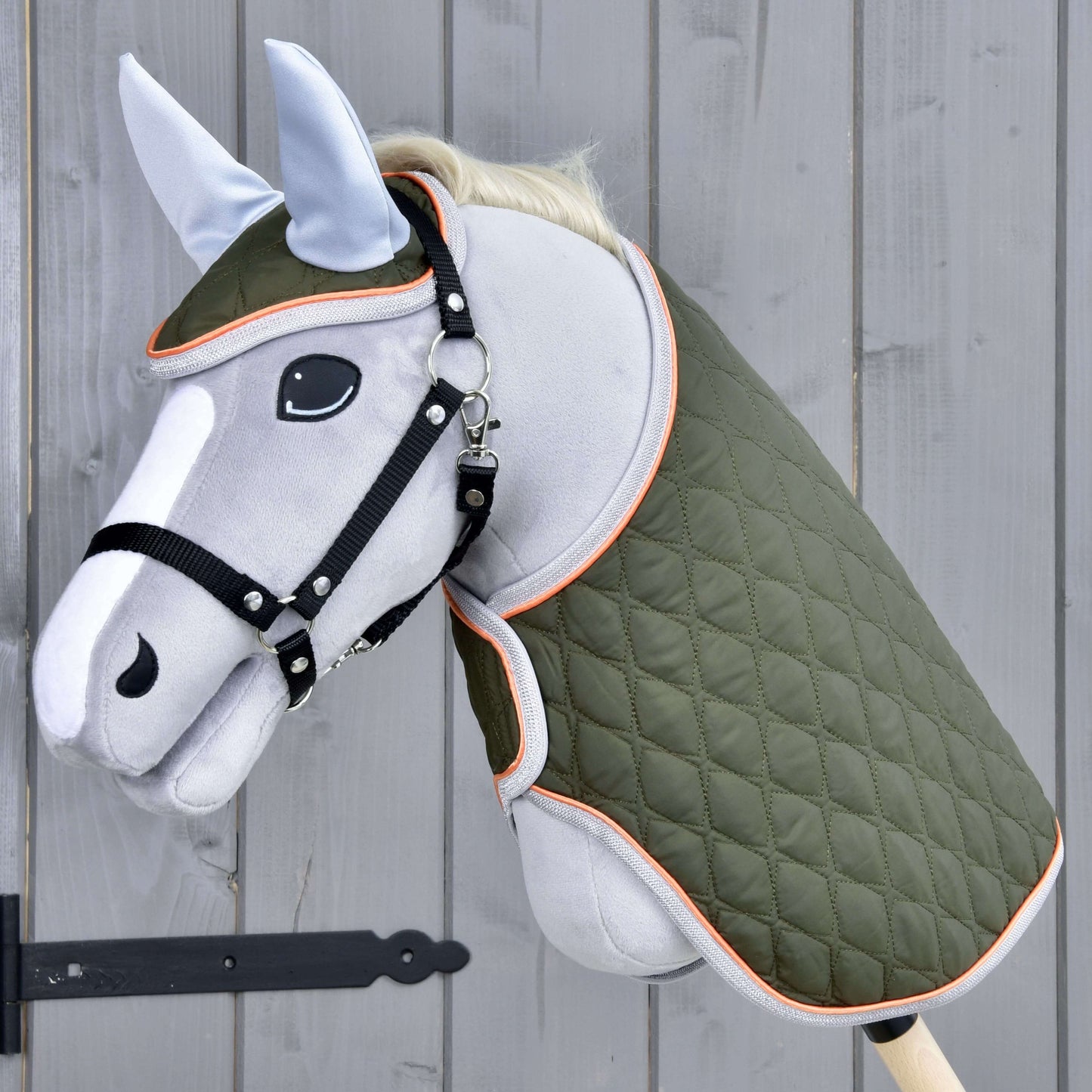 Hobby Horse LarDen Set Moos mit grauem Hobby Horse mit schwarzem Zaumzeug für Hobby Horse, grüner gesteppter Decke und hellblauer Fliegenhaube auf einem Holzstab, vor einer grauen Holzwand.