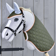 Hobby Horse LarDen Set Moos mit grauem Hobby Horse mit schwarzem Zaumzeug für Hobby Horse, grüner gesteppter Decke und hellblauer Fliegenhaube auf einem Holzstab, vor einer grauen Holzwand.