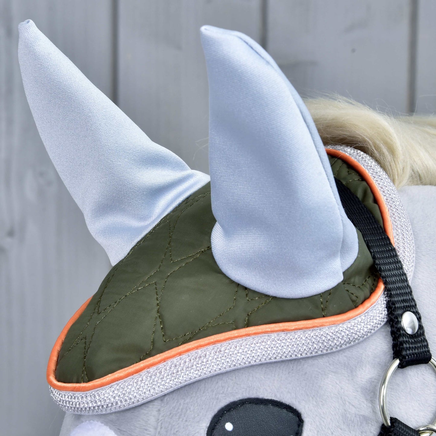 Nahaufnahme des Kopfes des Stoffpferdes Set Moos von Hobby Horse LarDen mit olivgrüner und orangefarbener Fliegenhaube, zwei hellgrauen spitzen Stoffohren und einer niedlichen Pferdedekoration vor hellgrauem Holz-Hintergrund.