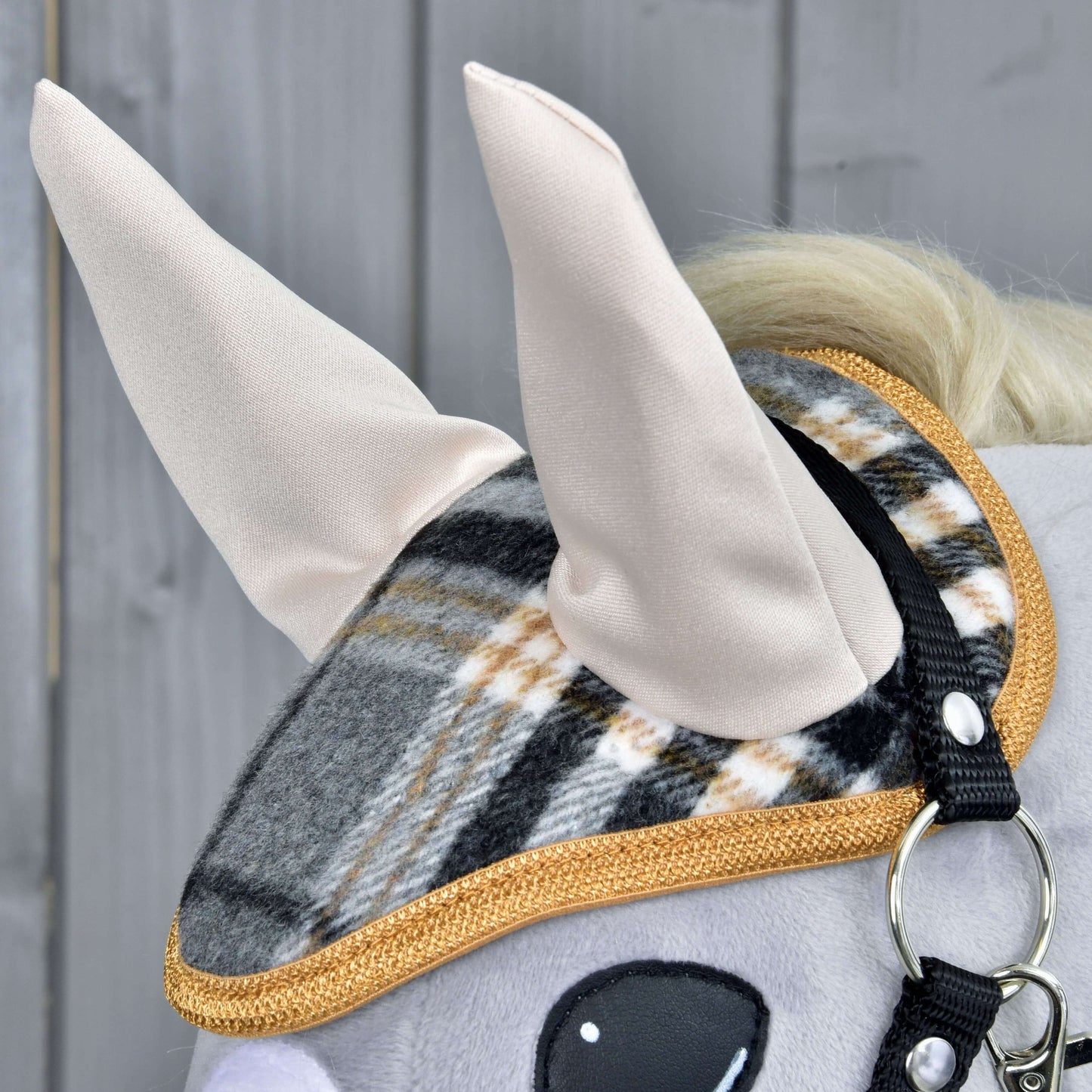 Close-up of the Hobby Horse LarDen "Set England Grey" plush horse head, featuring a plaid and gold-trimmed halter, ear bonnet, and soft fabric ears. Set against a gray wooden background—a charming horse decoration.