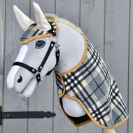 Hobby Horse LarDen Set England Grey mit grauem Plüsch-Hobby Horse mit schwarzem Zaumzeug für Hobby Horse und Zügeln sowie karierter Decke und passender Fliegenhaube, als charmante Dekoration vor Holz-Hintergrund.