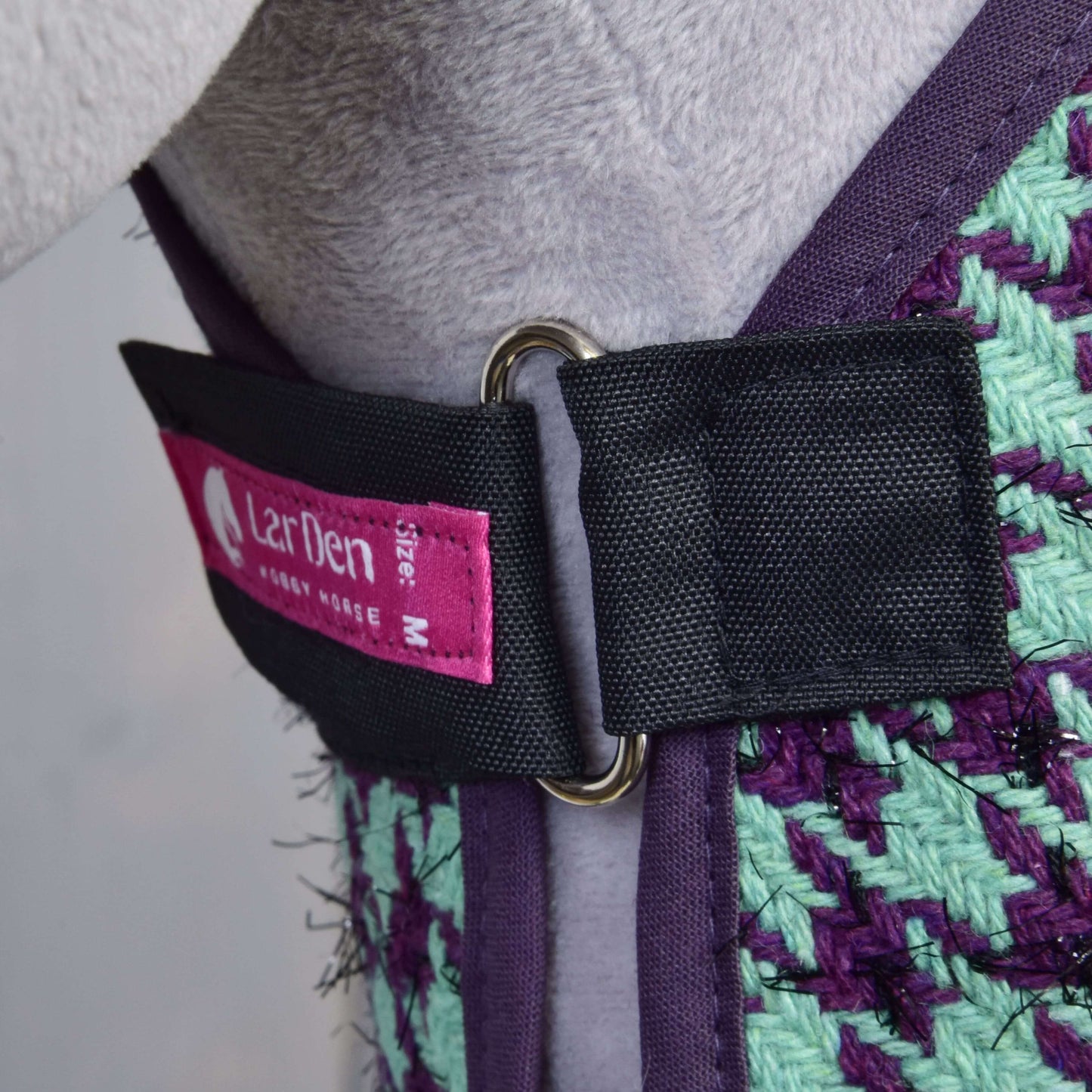 Nahaufnahme eines Geschirrs von Set Harmony Green von Hobby Horse LarDen in violett-grünem Gewebe, schwarzem Riemen mit Metall-D-Ring und rosa Größenetikett M. Getragen von einem hellgrauen Hobby Horse.