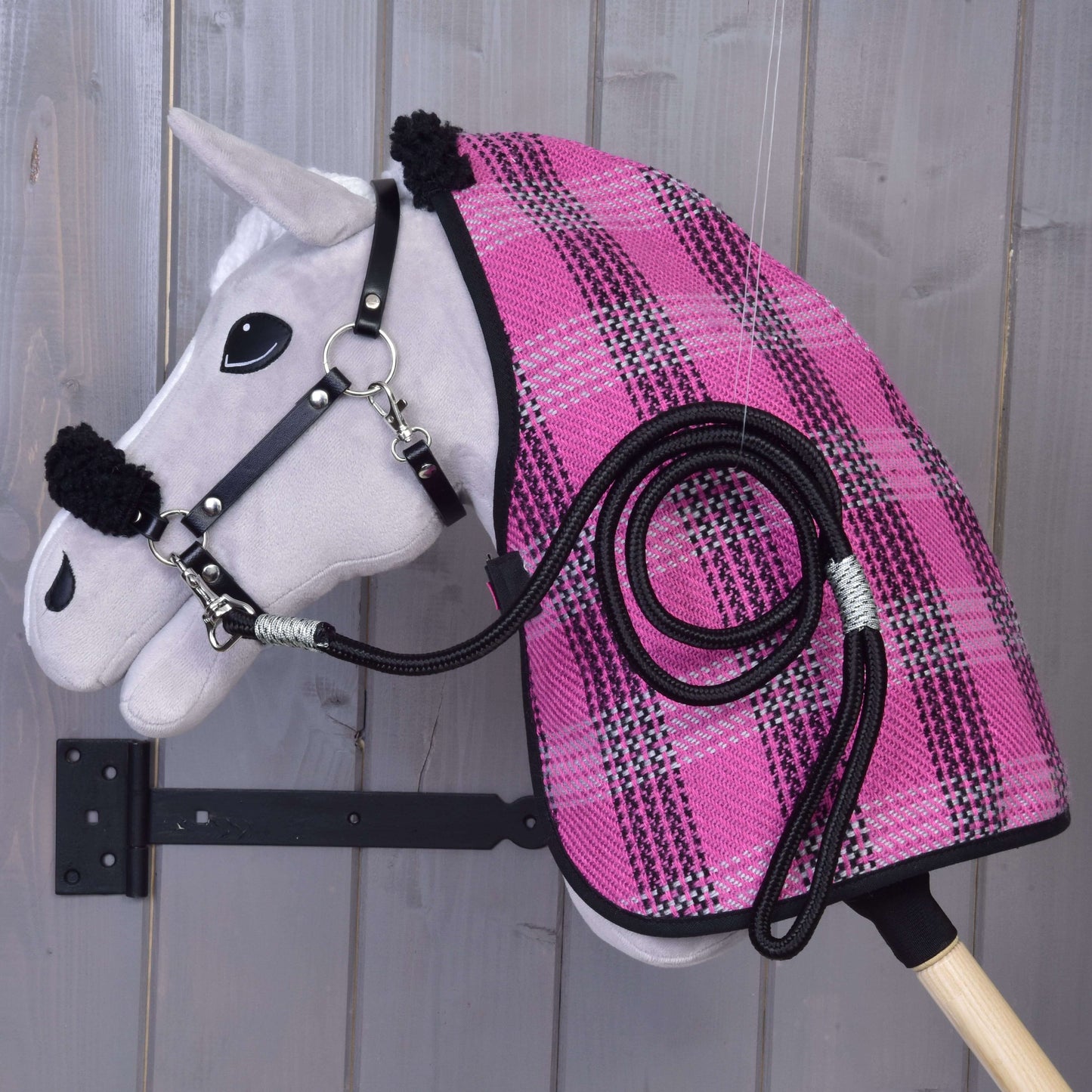 Ein graues Stickpferd Set Harmony Pink von Hobby Horse LarDen ist an einer Holzwand befestigt und mit einer pink-schwarz karierten Stalldecke, einem schwarzen Halfter und einem passenden aufgewickelten Führstrick an der Seite ausgestattet.