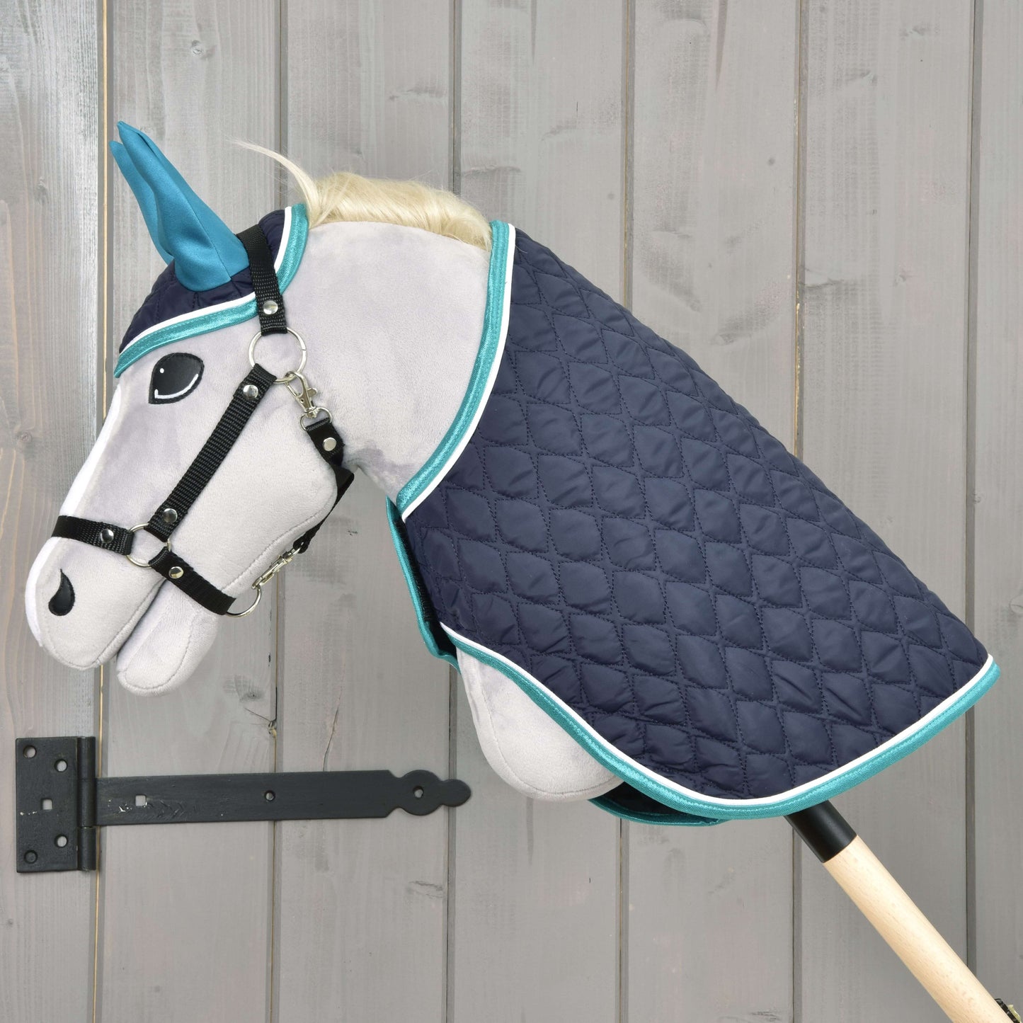 Ensemble Suprême Turquoise de Hobby Horse LarDen avec Hobby Horse en peluche gris, oreilles bleues, bride noire et couverture matelassée bleu marine à bordure turquoise, monté sur une perche en bois devant un mur en bois clair.