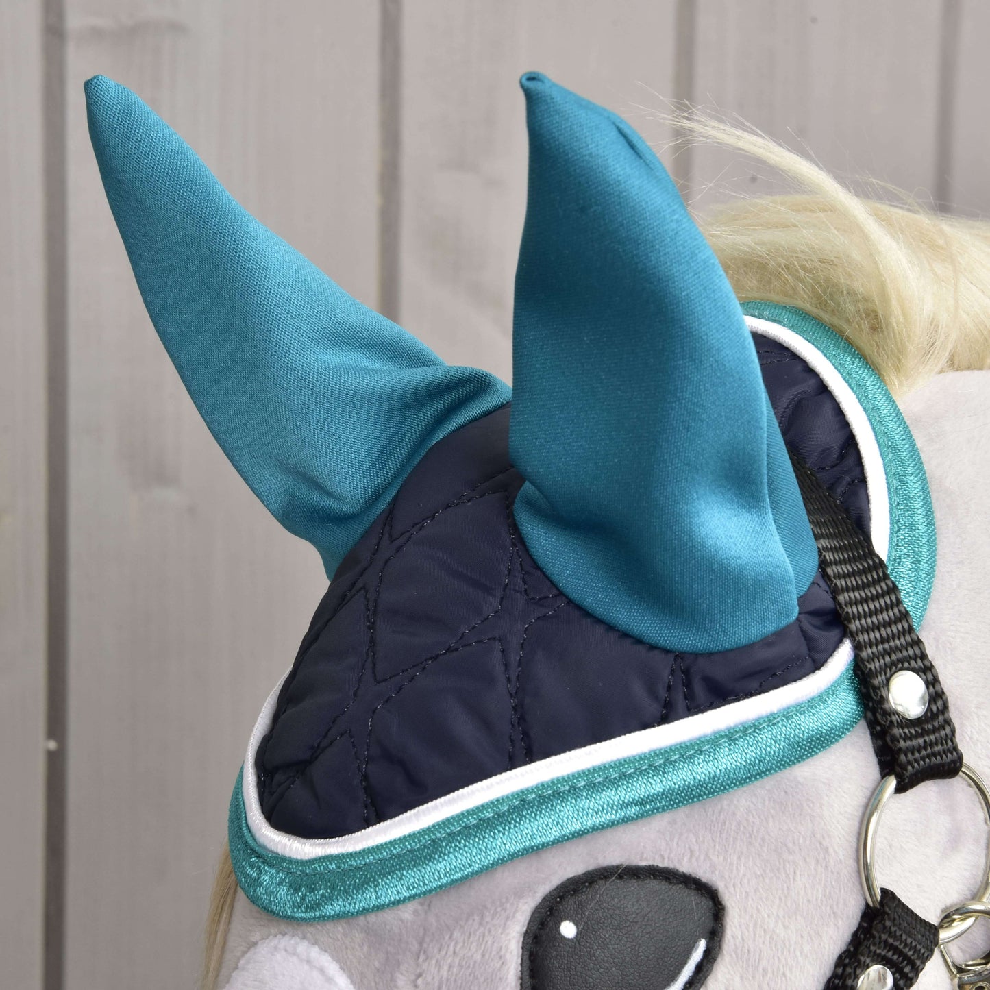 Gros plan sur l'Ensemble Suprême Turquoise de Hobby Horse LarDen : un cheval en peluche porte un masque pour les yeux matelassé bleu foncé avec protège-oreilles pointus turquoise et bordure turquoise, un ensemble de bonnet d'oreilles élégant, pour le jeu et la protection, sur fond de bois clair.