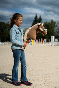 Lifestyle photo: a girl in a denim jacket stands on a sand riding arena holding the Hobby Horse Smart with Black Halter by Hobby Horse LarDen, jump poles behind - palomino plush head, long cream yarn mane - shown here with the black leatherette halter with complete fur.