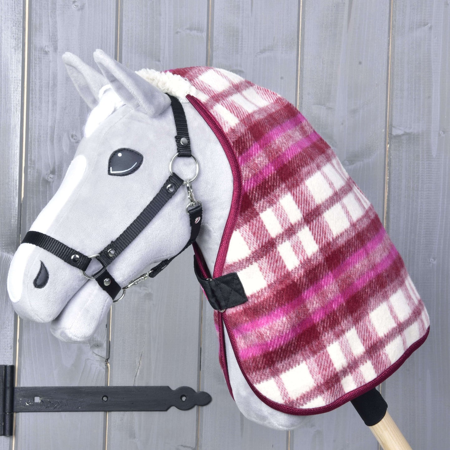Hobby Horse gris avec bride noire portant la Couverture d'écurie LarDen rouge et blanche à carreaux de Hobby Horse LarDen autour de l’encolure. Le fond montre un mur en bois à planches verticales.