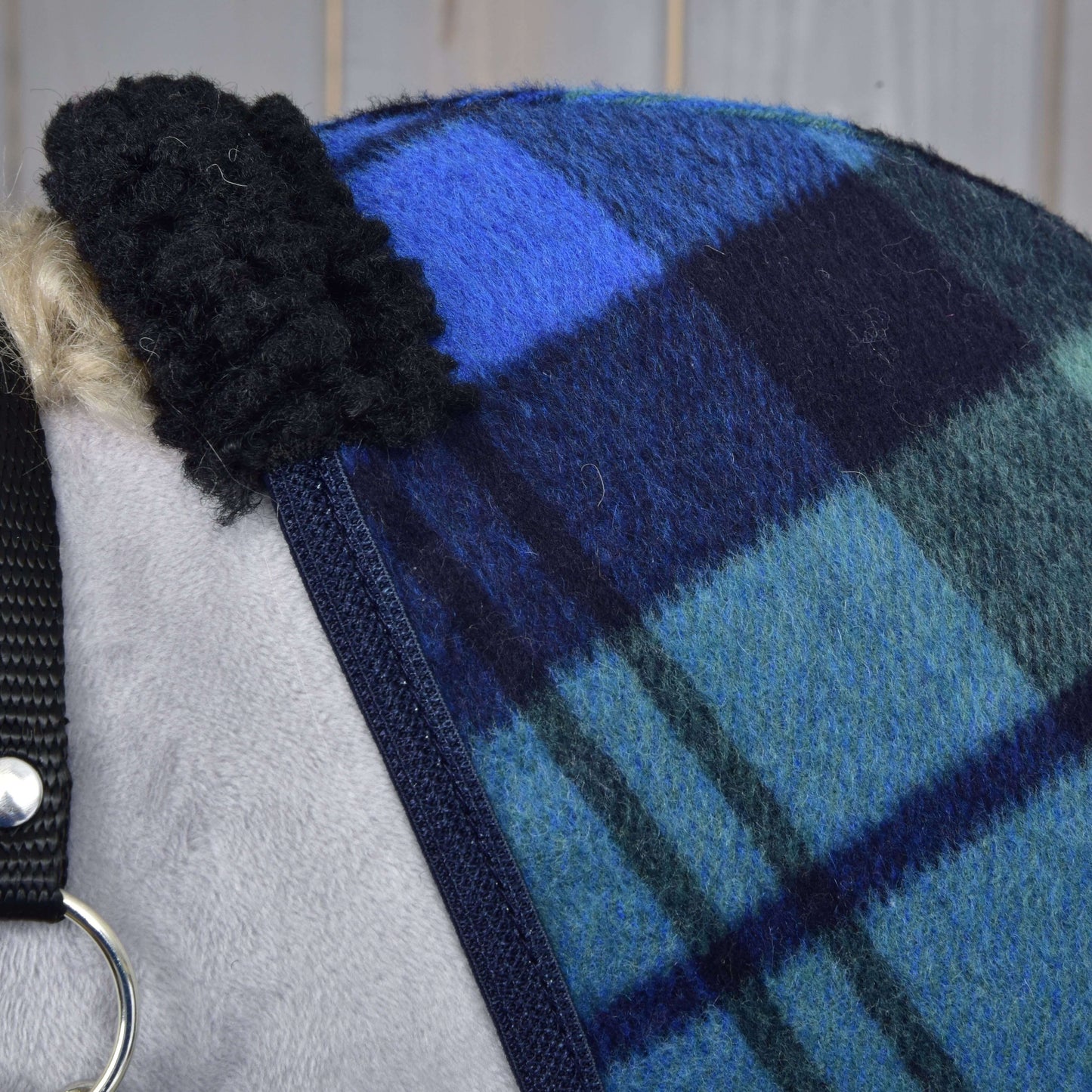 Gros plan sur la Couverture d'écurie Perth de Hobby Horse LarDen, avec motif en laine à carreaux bleus et verts et bordure noire duveteuse, drapée sur un cheval. Une sangle en nylon noire et un anneau métallique sont visibles à gauche.