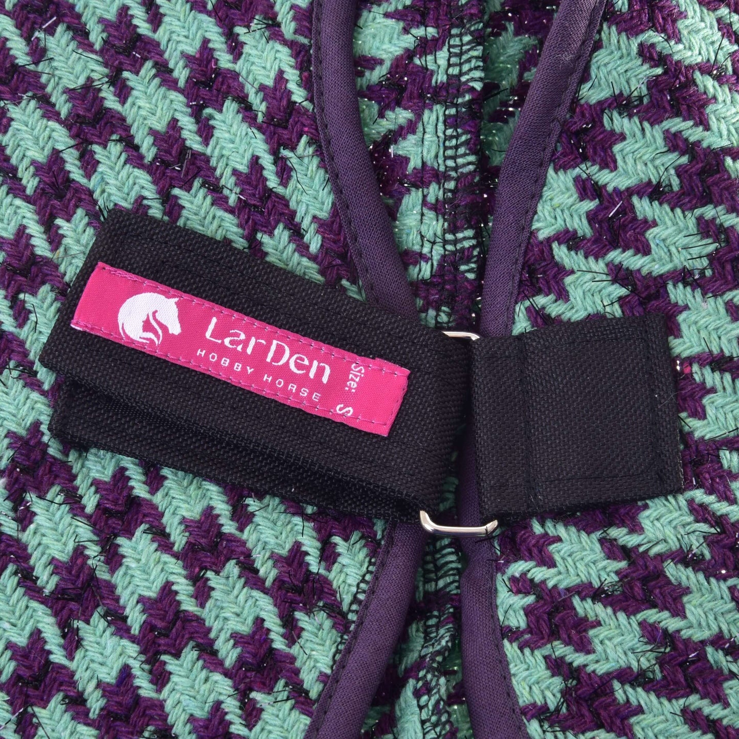 Gros plan sur la Couverture d'écurie verte de Hobby Horse LarDen, montrant un tissu tissé vert et violet en zigzag avec sangle noire et étiquette rose avec logo cheval blanc, parfaite comme accessoire élégant pour votre Hobby Horse.