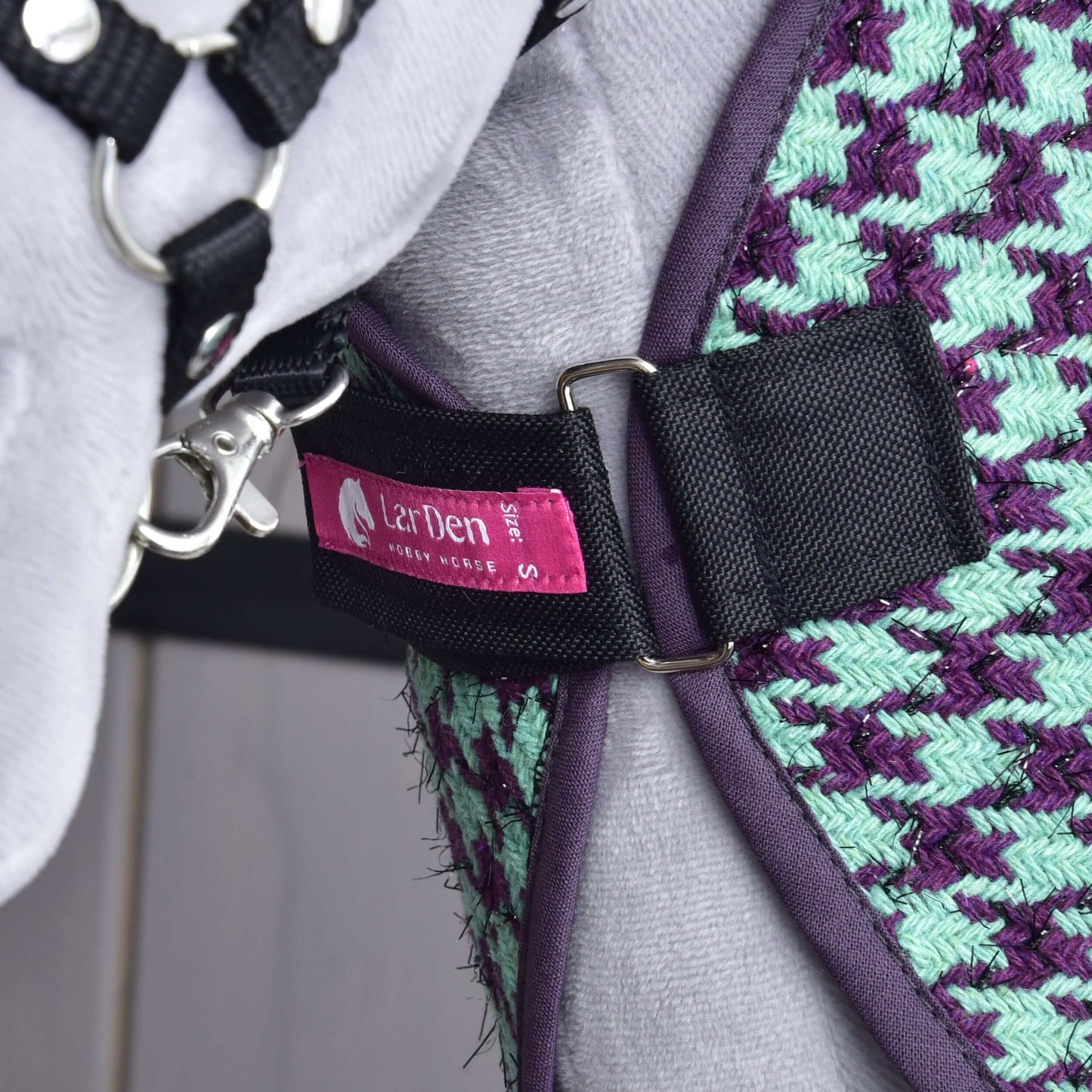 Gros plan sur un cheval portant la Couverture d'écurie verte de Hobby Horse LarDen, avec tissu tissé vert et violet, sangle noire, étiquette rose « LarDen » et mousqueton en métal attaché.