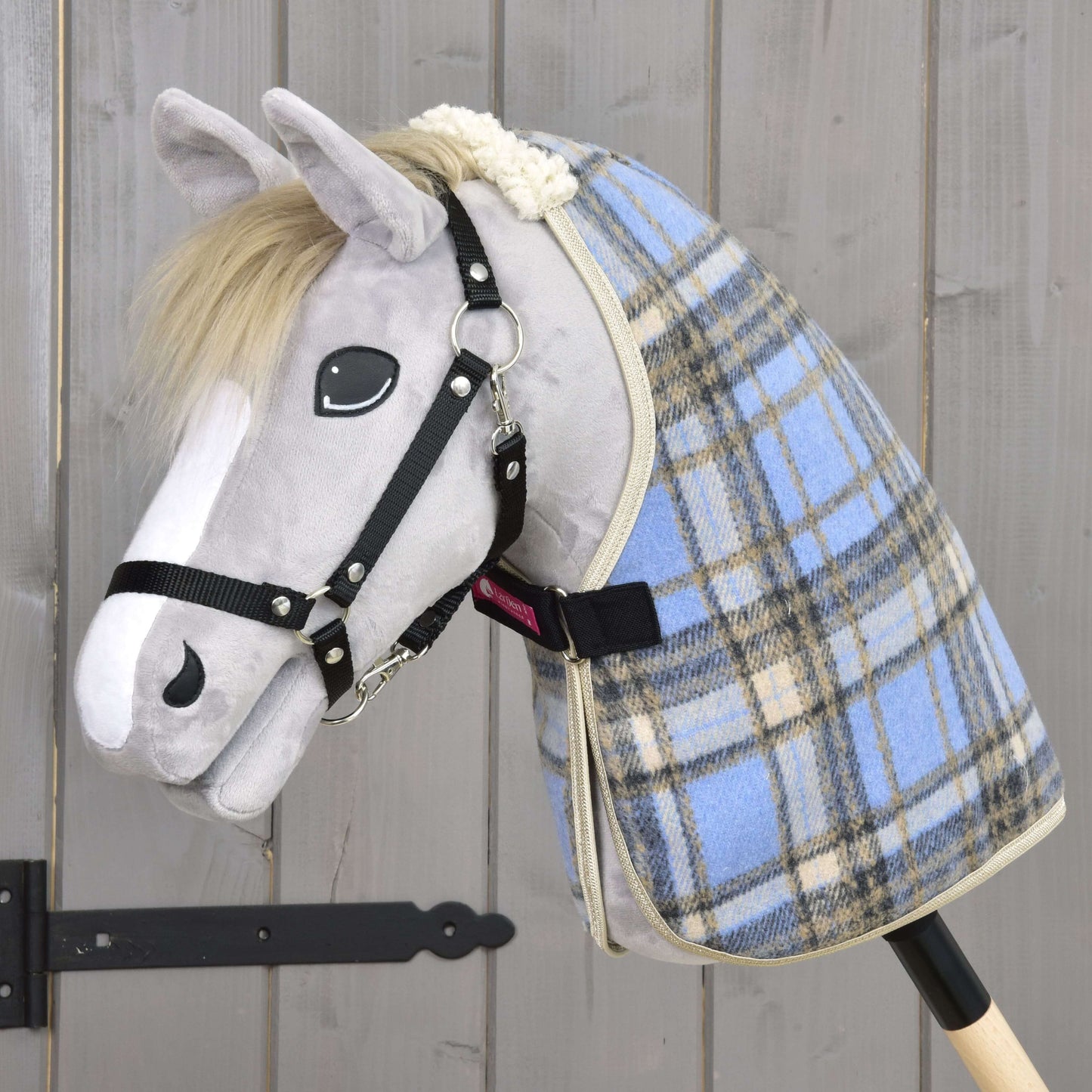 Ein Plüsch-Hobby Horse mit grauem Gesicht, weißer Mähne und schwarzem Zaumzeug trägt die Stalldecke Light Blue von Hobby Horse LarDen, ist auf einem Holzstab montiert und steht vor einer rustikalen Holzwand.