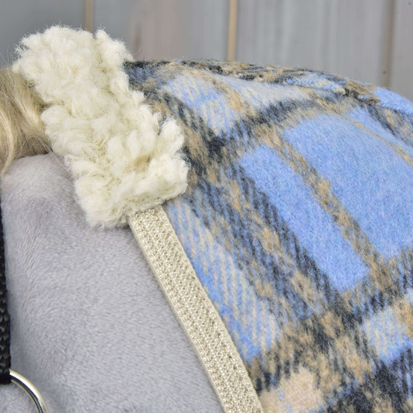 Nahaufnahme der Stalldecke Light Blue von Hobby Horse LarDen mit blauem, beigem und grauem Karomuster und cremefarbenem Sherpa-Besatz, über eine graue Oberfläche drapiert. Links sind teilweise ein schwarzer Riemen und ein silberner Ring sichtbar.