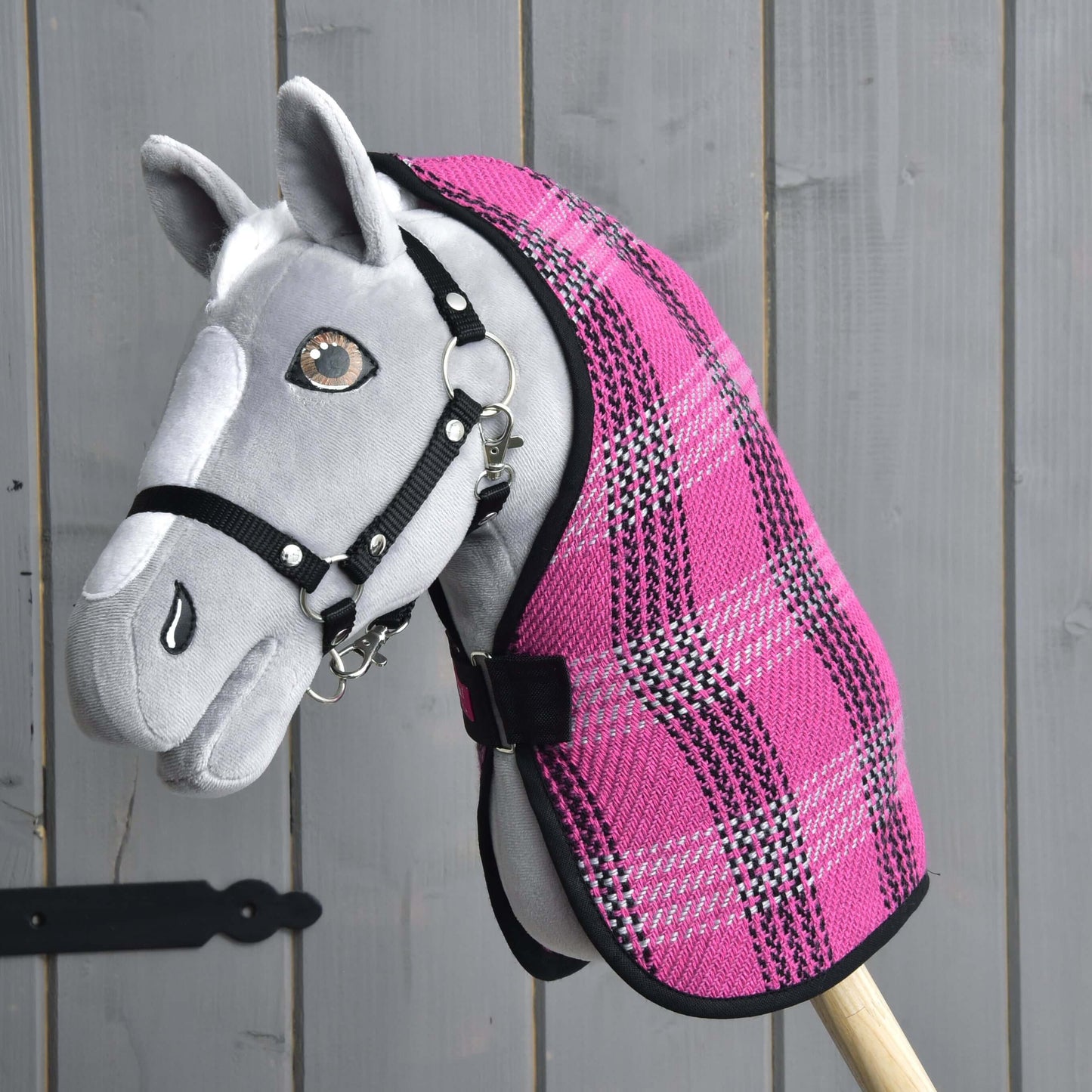 Hobby Horse gris de Hobby Horse LarDen avec bride noire et Couverture d'écurie rose, monté sur un bâton en bois et présenté devant un mur en bois gris.