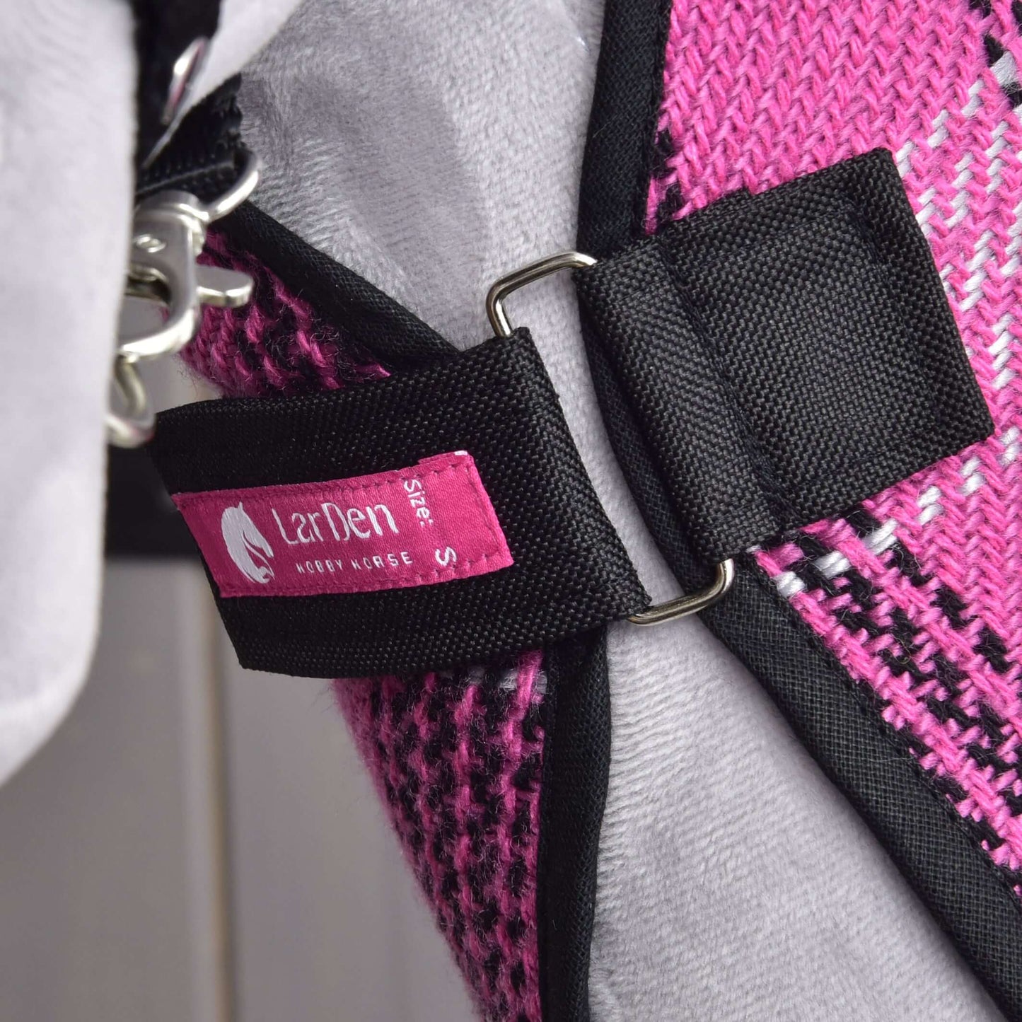 Gros plan sur un harnais en tissu rose et noir avec anneau en métal et étiquette Hobby Horse LarDen Stable Blanket Pink Size S, attaché à une matière grise, possiblement une partie d'une couverture d'entraînement pour cheval ou pour Hobby Horse, portée par un animal de compagnie.