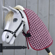 A gray hobby horse with a black bridle wears the pink houndstooth-patterned Stable Blanket Winter Magic by Hobby Horse LarDen, made of soft PES, posed before a light gray wooden background.