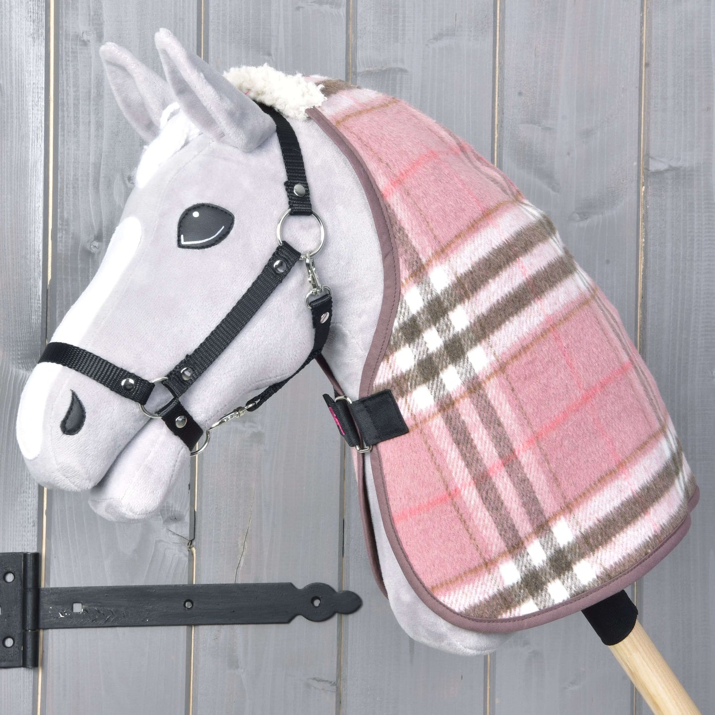 Die Hobby Horse LarDen „Stalldecke Lady“ ist ein graues Plüsch-Hobby Horse mit einer rosa-braun karierten Decke und schwarzem Zaumzeug auf einem Holzstab vor grauem Holz-Hintergrund.