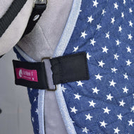Close-up of the blue quilted Stable Blanket Star by Hobby Horse LarDen, featuring white stars, a black strap with silver buckle, and a pink Izefan label, worn by a gray animal as part of its harness or vest.