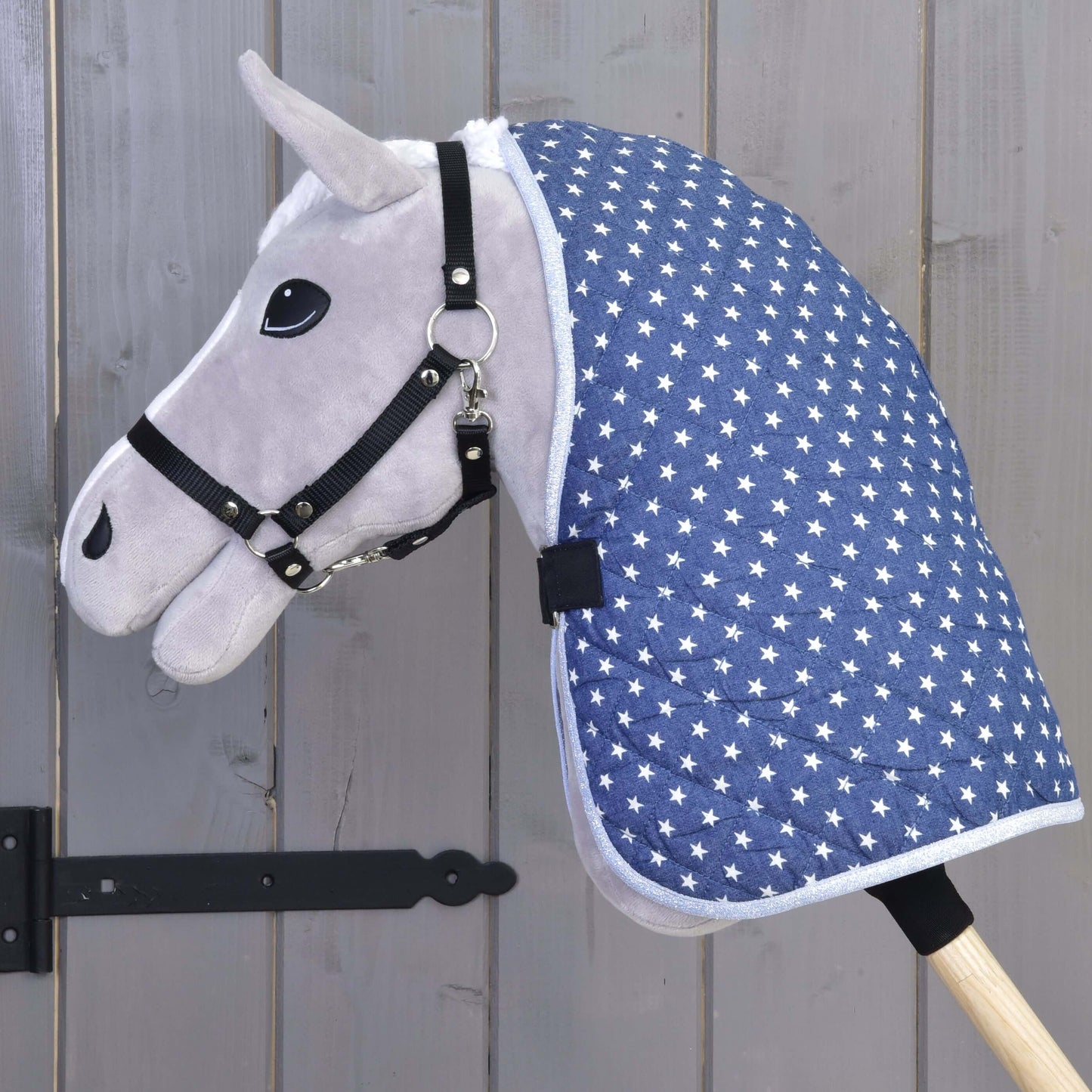 Jouet Hobby Horse gris de Hobby Horse LarDen, avec bride noire et Couverture d'écurie Étoile, monté sur un bâton en bois et présenté devant une porte en bois grise.