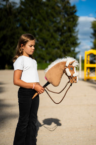 Mädchen in weißem Oberteil führt das Hobby Horse Star mit schwarzem Halfter von Hobby Horse LarDen über einen Sandreitplatz, dahinter ein gelber Hindernisständer – Palomino-Plüschkopf, wehende weiße Garnmähne – mit braunem Halfter mit vollständigem Fellbesatz und Vivaldi-Zügeln