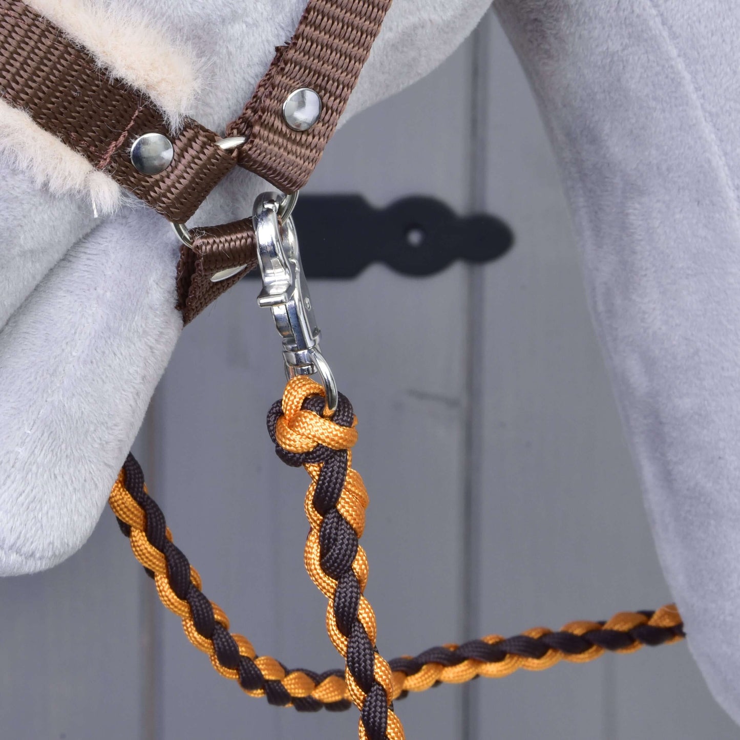 Gros plan sur une tête de cheval portant l’Ensemble Combinaison Parfaite Marron de Hobby Horse LarDen, avec un licol rembourré de fourrure et une longe tressée orange-marron avec mousqueton argenté. Porte grise floue avec plaque de serrure noire en arrière-plan, idéal pour les fans de Hobby Horse.