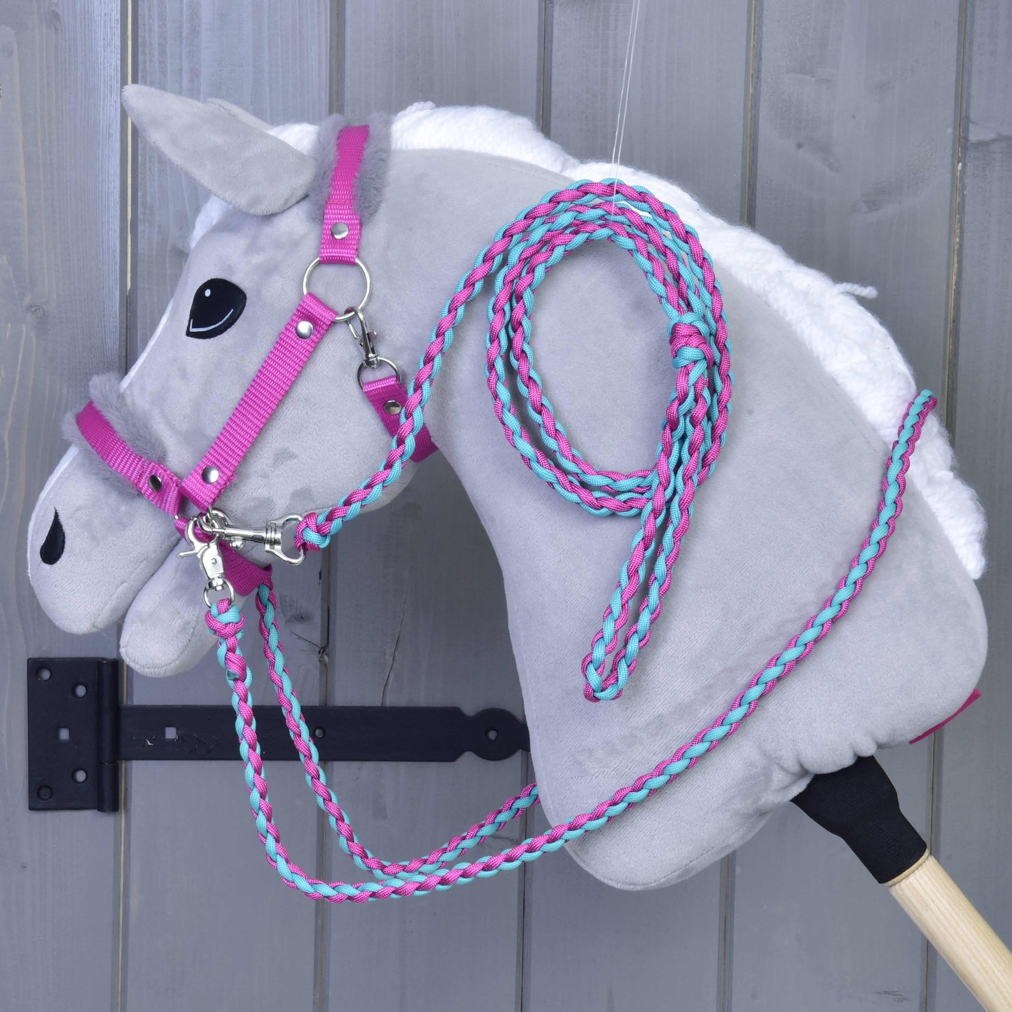 Ensemble Combinaison Parfaite Rose de Hobby Horse LarDen sur un Hobby Horse en peluche gris avec licol et rênes rose et turquoise rembourrés de fourrure, suspendu à un crochet mural sur fond de bois gris.