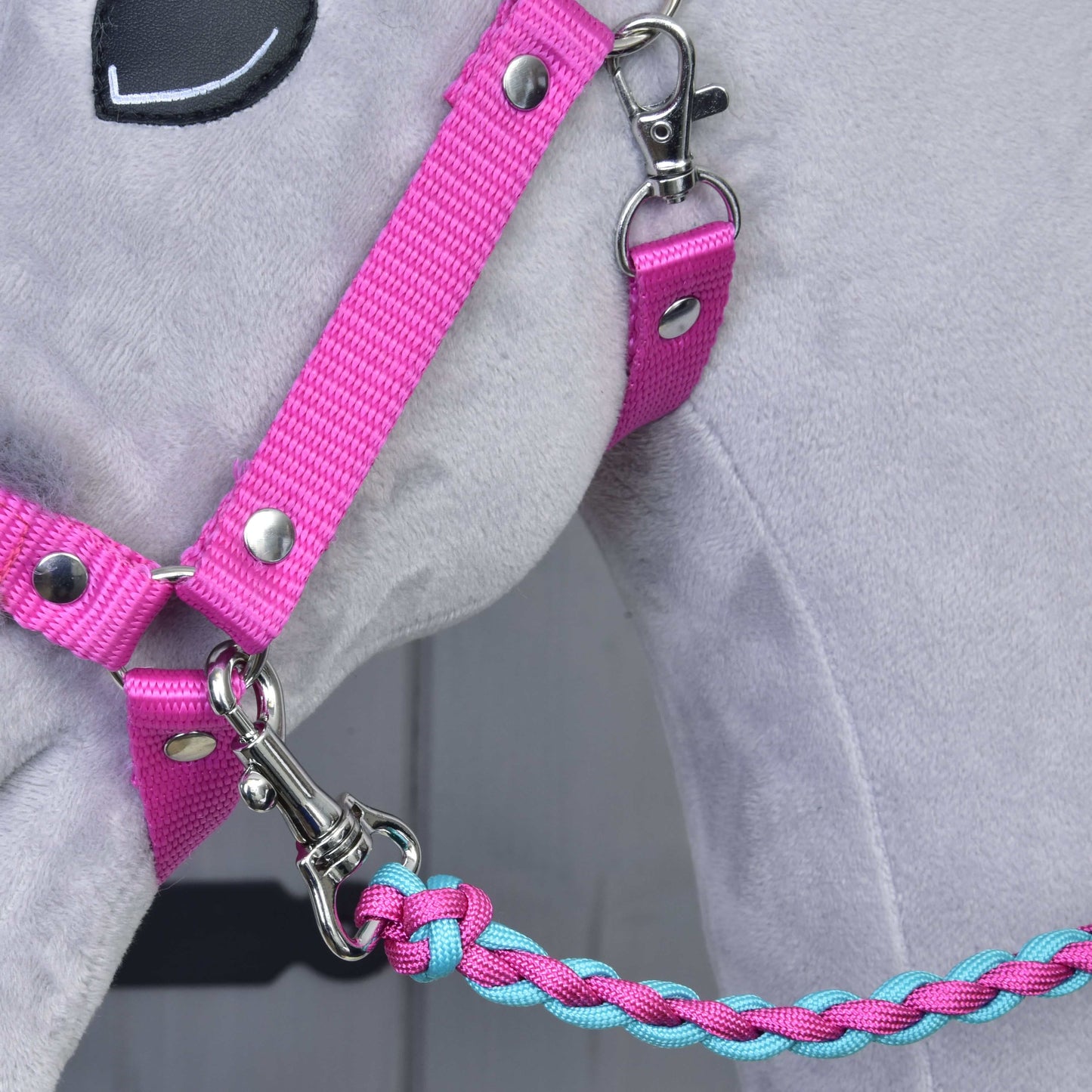Gros plan sur un cheval en peluche gris portant l’Ensemble Combinaison Parfaite Rose de Hobby Horse LarDen : un licol rose rembourré de fourrure avec une longe tressée bleue et rose attachée par un mousqueton argenté, idéal pour les amateurs d’accessoires pour Hobby Horse.