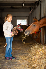 Mädchen in cremefarbener Jacke hält das Hobby Horse Stormy mit schwarzem Halfter von Hobby Horse LarDen zu einem echten braunen Pferd, das in einem Stall Heu frisst – brauner Plüschkopf mit Apfelschimmelung, schwarze Garnmähne – mit Burgundy Fellhalfter aus Kunstleder