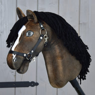 Hobby Horse Sundance mit schwarzem Halfter von Hobby Horse LarDen mit braunem Kopf, inspiriert von der Schnelligkeit von Hengsten, schwarzer Garnmähne, weißer Blesse und schwarzem Halfter, auf einem stabilen Holzstab vor einer grauen Holzwand.