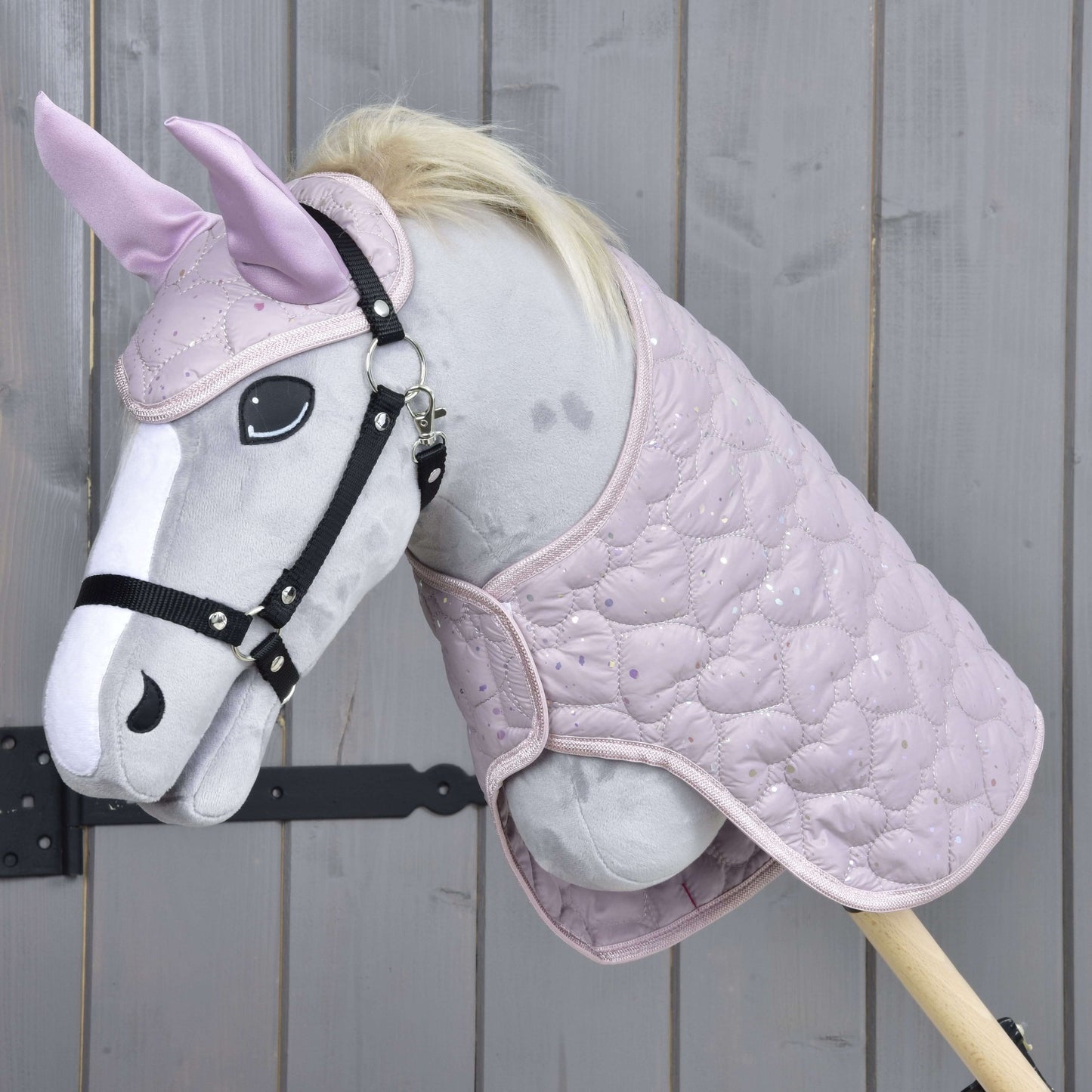 Das Set Sweetest Heart von Hobby Horse LarDen zeigt ein Plüsch-Hobby Horse mit hellgrauem Gesicht, schwarzem Zaumzeug, heller Mähne, rosa Ohren mit Fliegenhaube und gesteppter rosa Decke, vor einem Holz-Hintergrund.