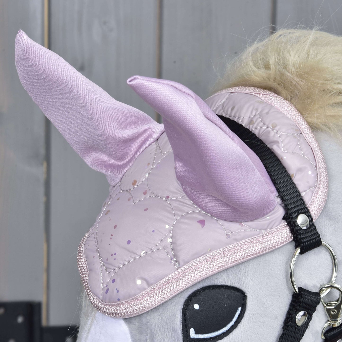 Eine Nahaufnahme von Set Sweetest Heart von Hobby Horse LarDen zeigt den hellgrauen Kopf mit einem glänzenden gesteppten rosa Einhornhorn, einer Fliegenhaube, schwarzem Zaumzeug und einem Holz-Hintergrund.