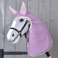 Die Hobby Horse LarDen „Regendecke Sweetheart“ ist ein Plüsch-Hobby Horse mit grauem Gesicht, schwarzem Zaumzeug, rosa Ohren und lavendelfarbener Regendecke mit Herzmuster – perfekt für jedes Wetter – auf einem Holzstab vor einer Holzwand.
