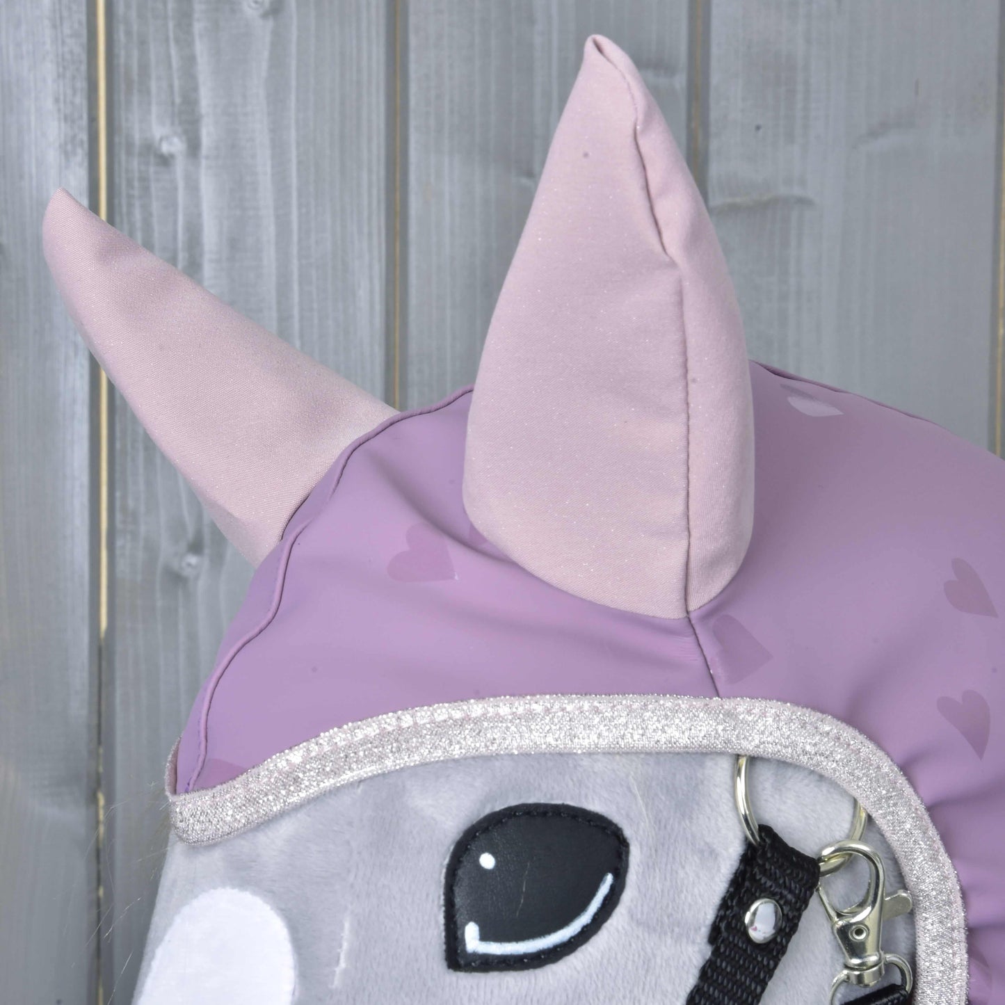 Gros plan sur la tête de cheval en peluche « Imperméable Chéri » de Hobby Horse LarDen, avec oreilles violet clair et housse à motif cœur, sur fond en bois, charmant accessoire tout temps pour Hobby Horse.